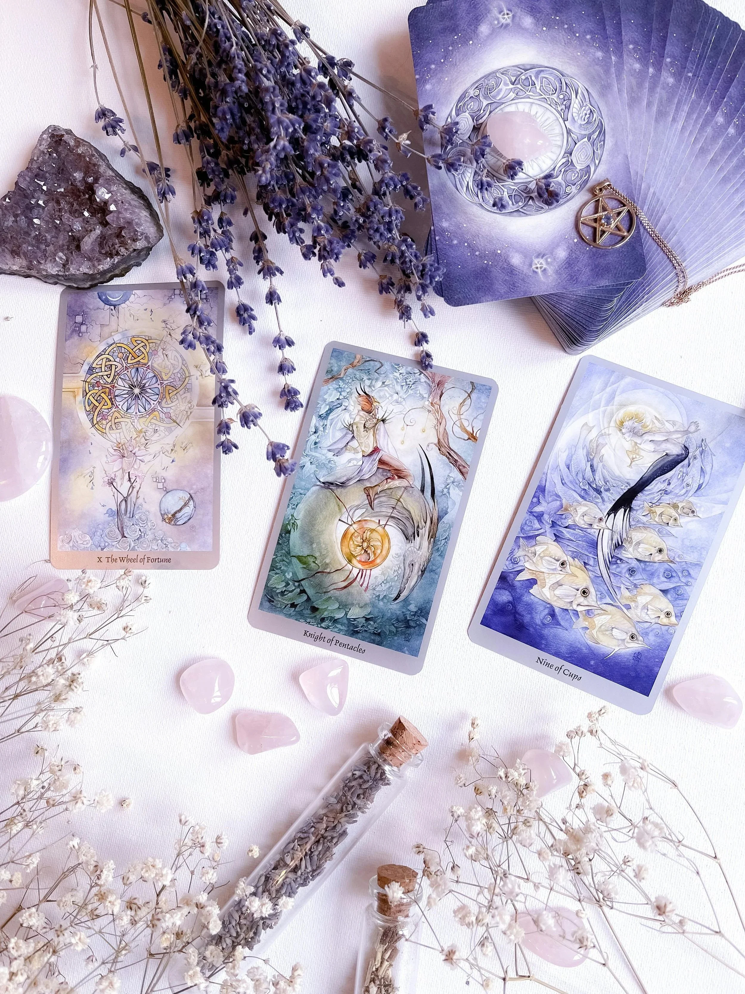 An arrangement of tarot and oracle cards on a white surface, with dried lavender flowers, rose quartz stones, a large clear quartz crystal, and tiny glass vials of dried herbs. The cards include the Wheel of Fortune, Knight of Pentacles, and Nine of Cups, with a tarot deck in a purple box and a pentacle pendant nearby.