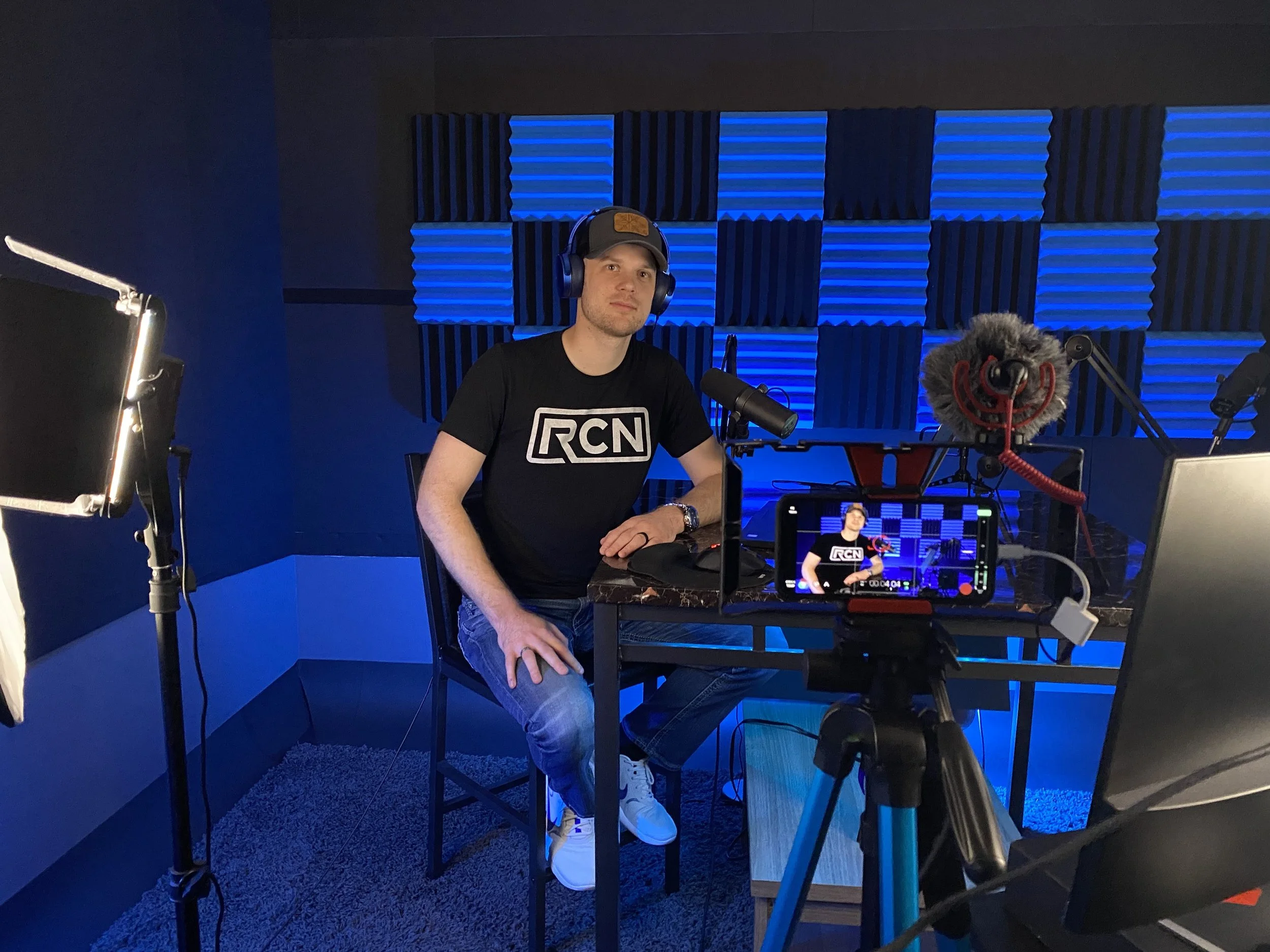 Man sitting at a desk with podcasting or recording equipment, including microphone, camera, headphones, and soundproofed blue and black acoustic panels in the background.