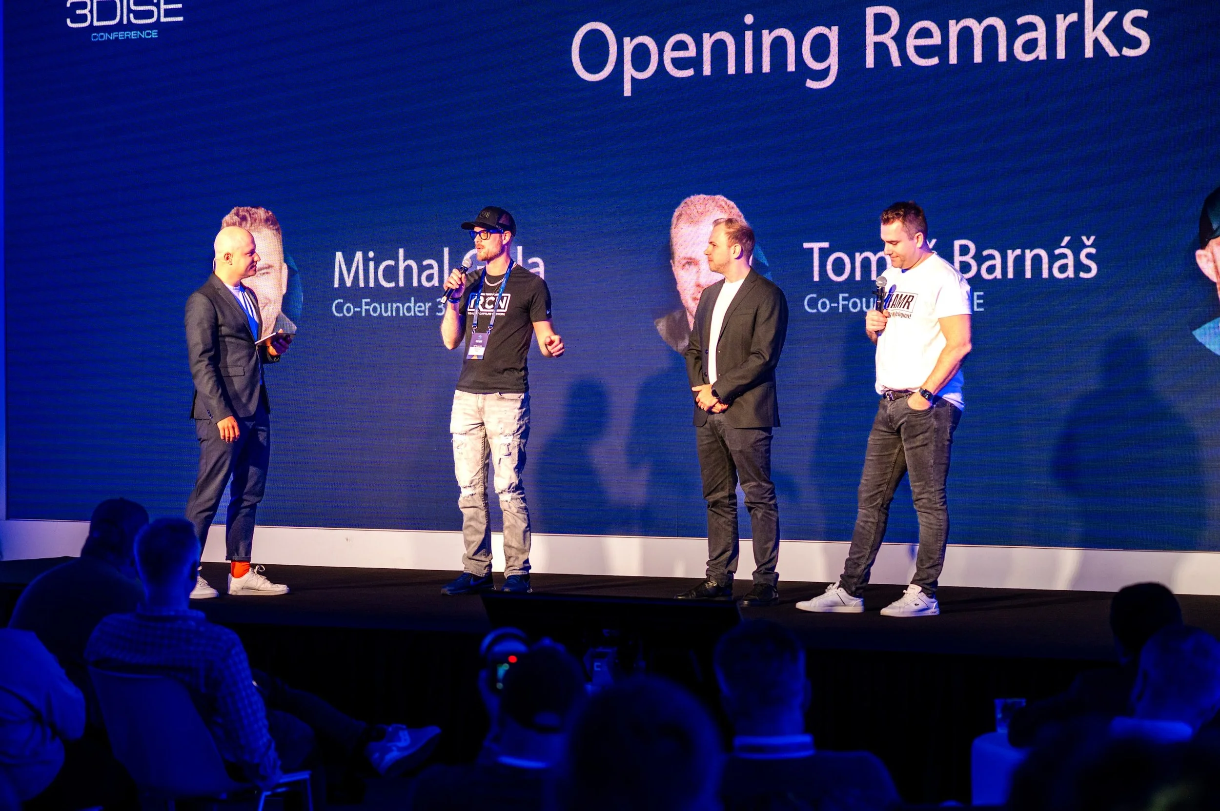 Four men on stage during a conference presentation, with a large screen behind them displaying the text 'Opening Remarks'. The man on the far left is holding a microphone and a tablet, and the three others are standing with microphones. The audience is visible in the foreground.