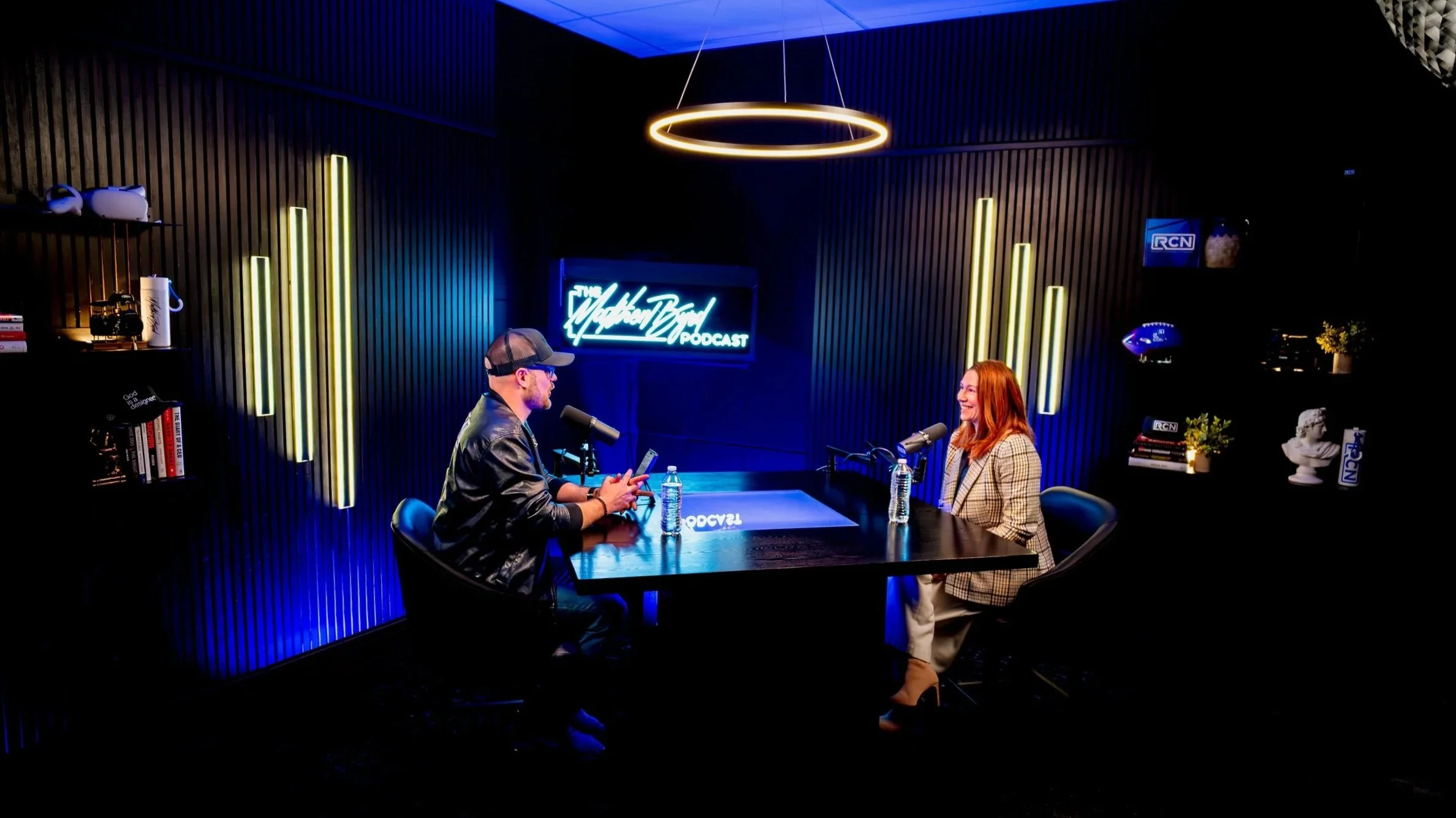 Two people a man and a woman having a conversation in a podcast studio with dark wooden walls, neon signs, and shelves with books and decorations.
