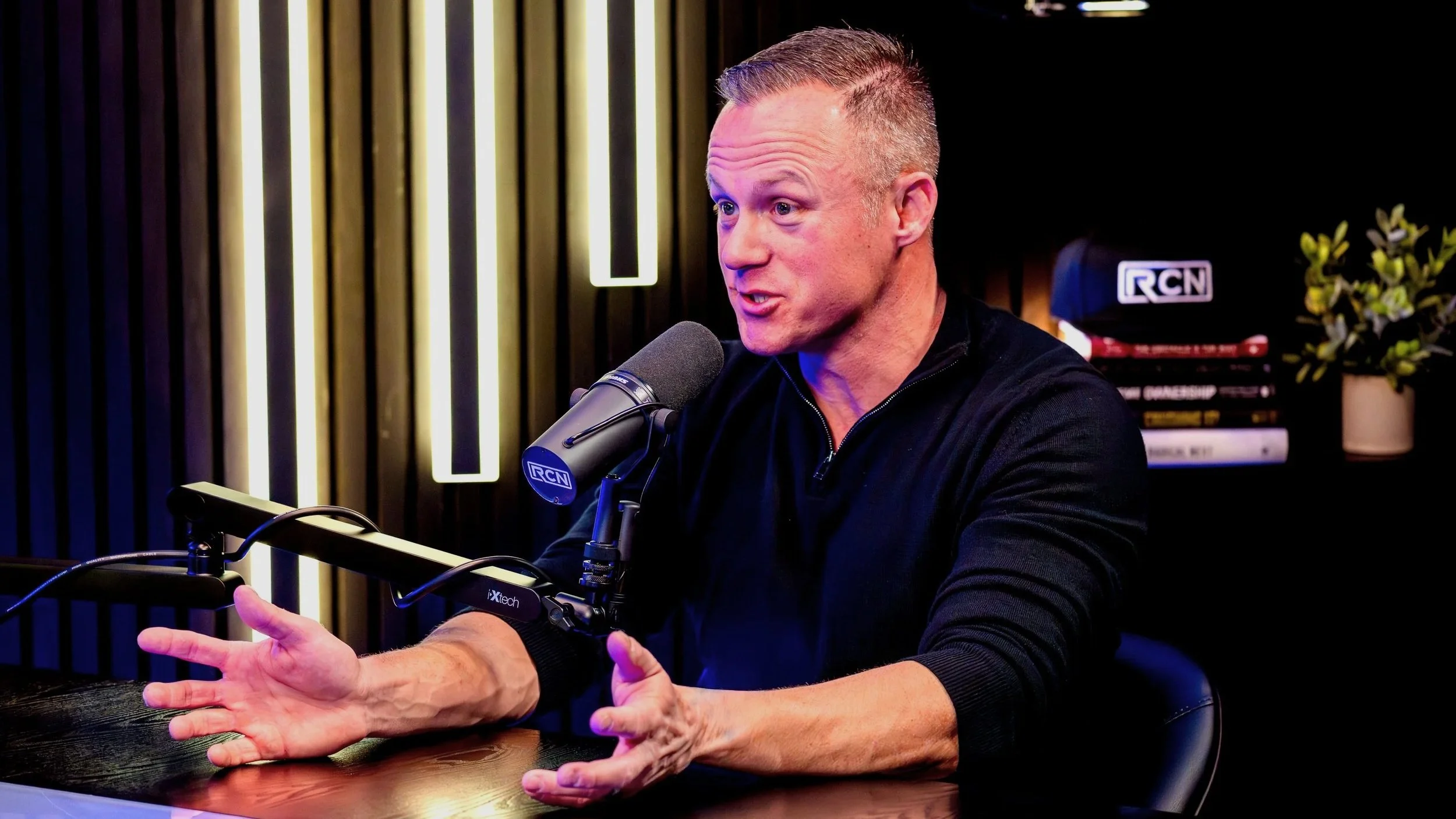 A man with short gray hair speaking into a microphone during a podcast or interview, gesturing with his hands, in a modern studio setting with books and a potted plant in the background.