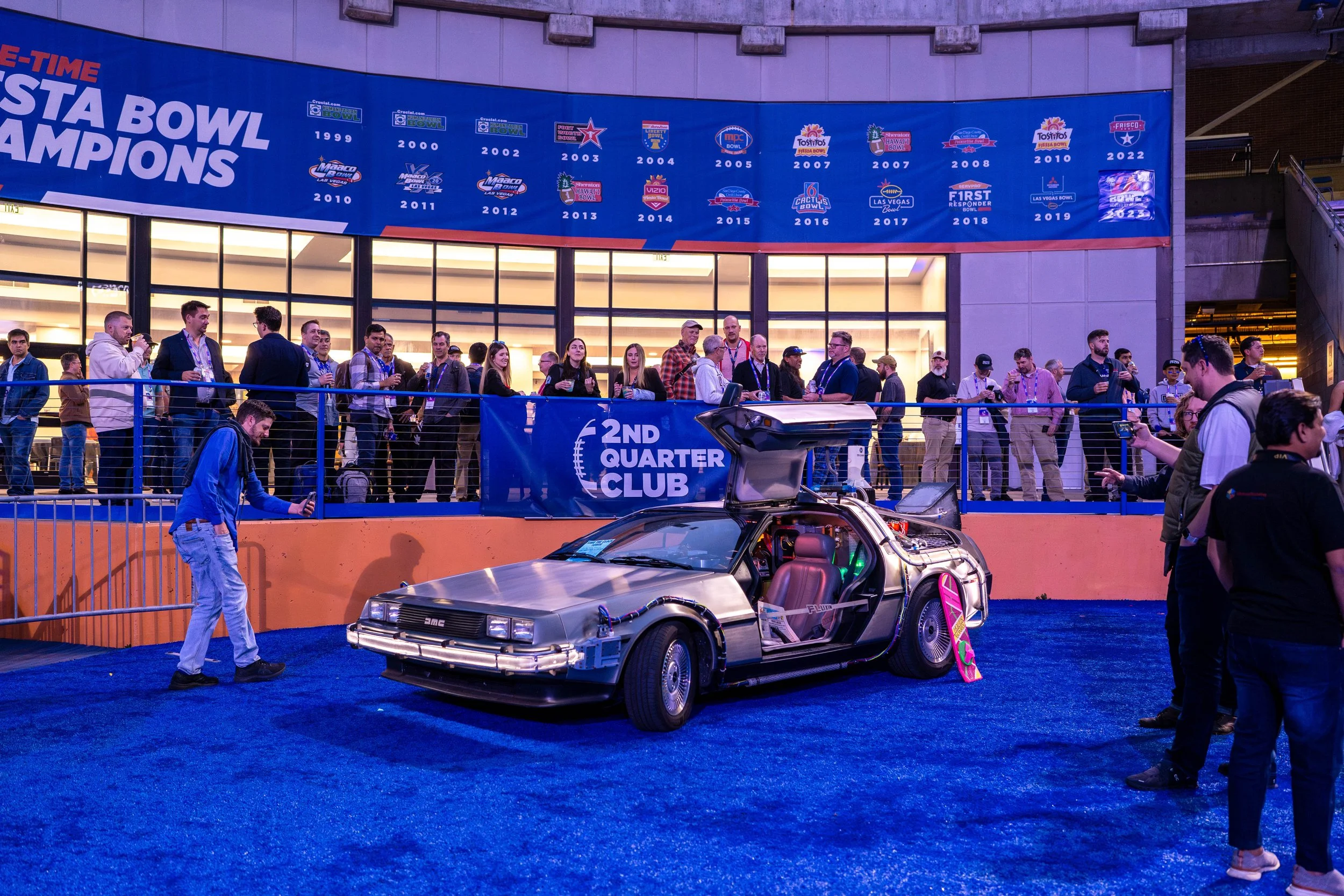 A DeLorean time machine from the movie 'Back to the Future' displayed at an event, with a crowd of people around and a large banner showing several sports team championships above.
