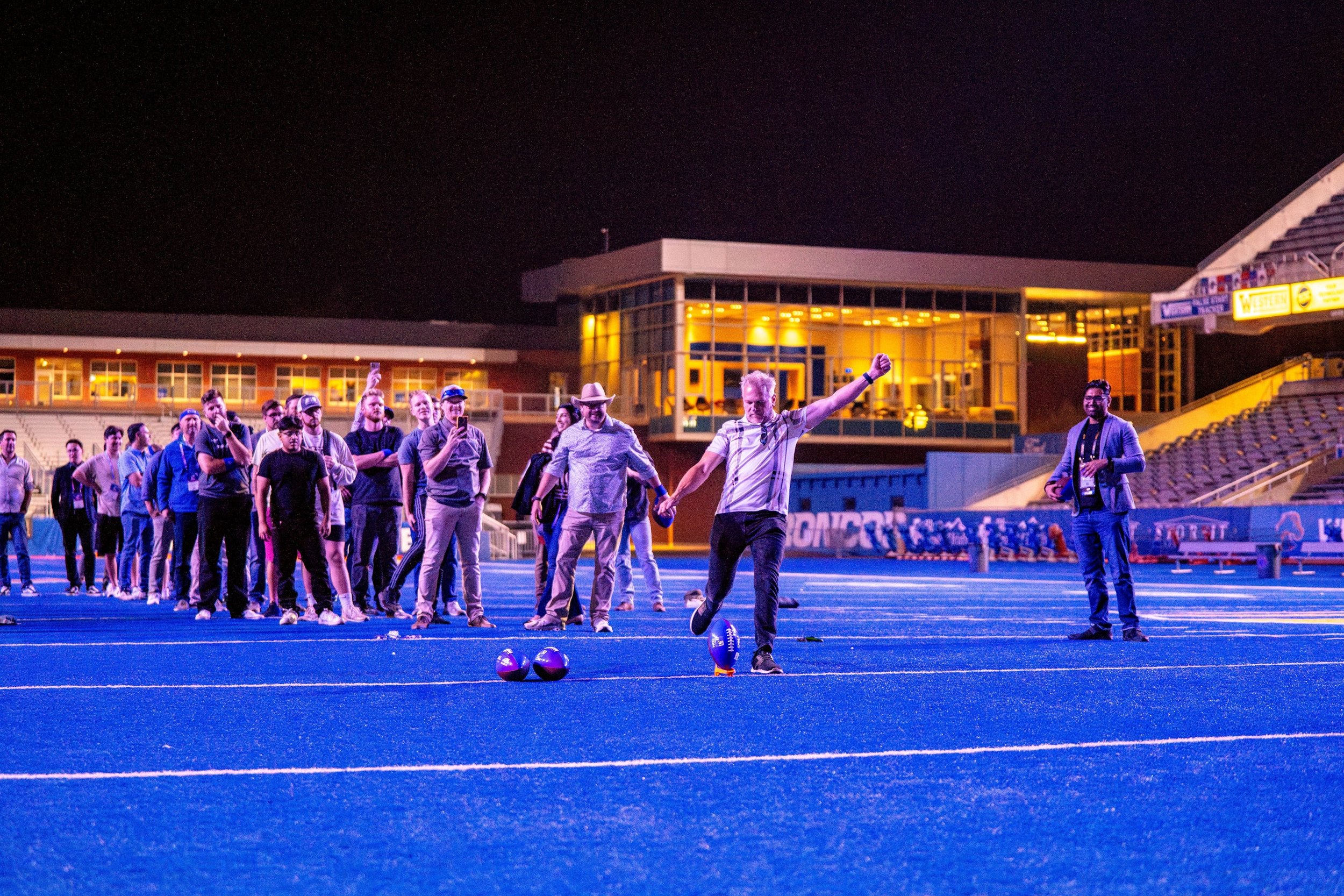 A group of people on a sports field at night, with some holding hands, and two with footballs at their feet, in front of a modern building with large glass windows and bright yellow lights.