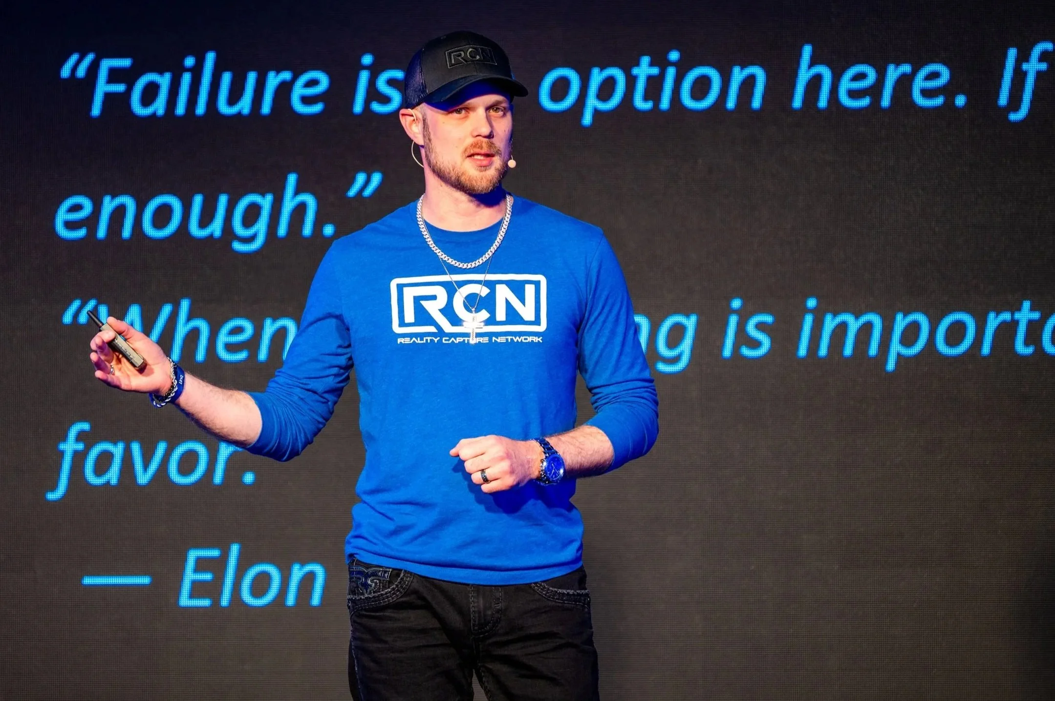Elon Musk giving a presentation, standing in front of a large screen displaying a quote about failure, wearing a blue RCN shirt, black cap, and jewelry.