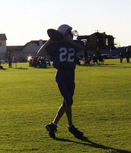 A football player in a blue uniform with the number 22 on it, holding a football on his shoulder during a game on a grassy field at sunset.