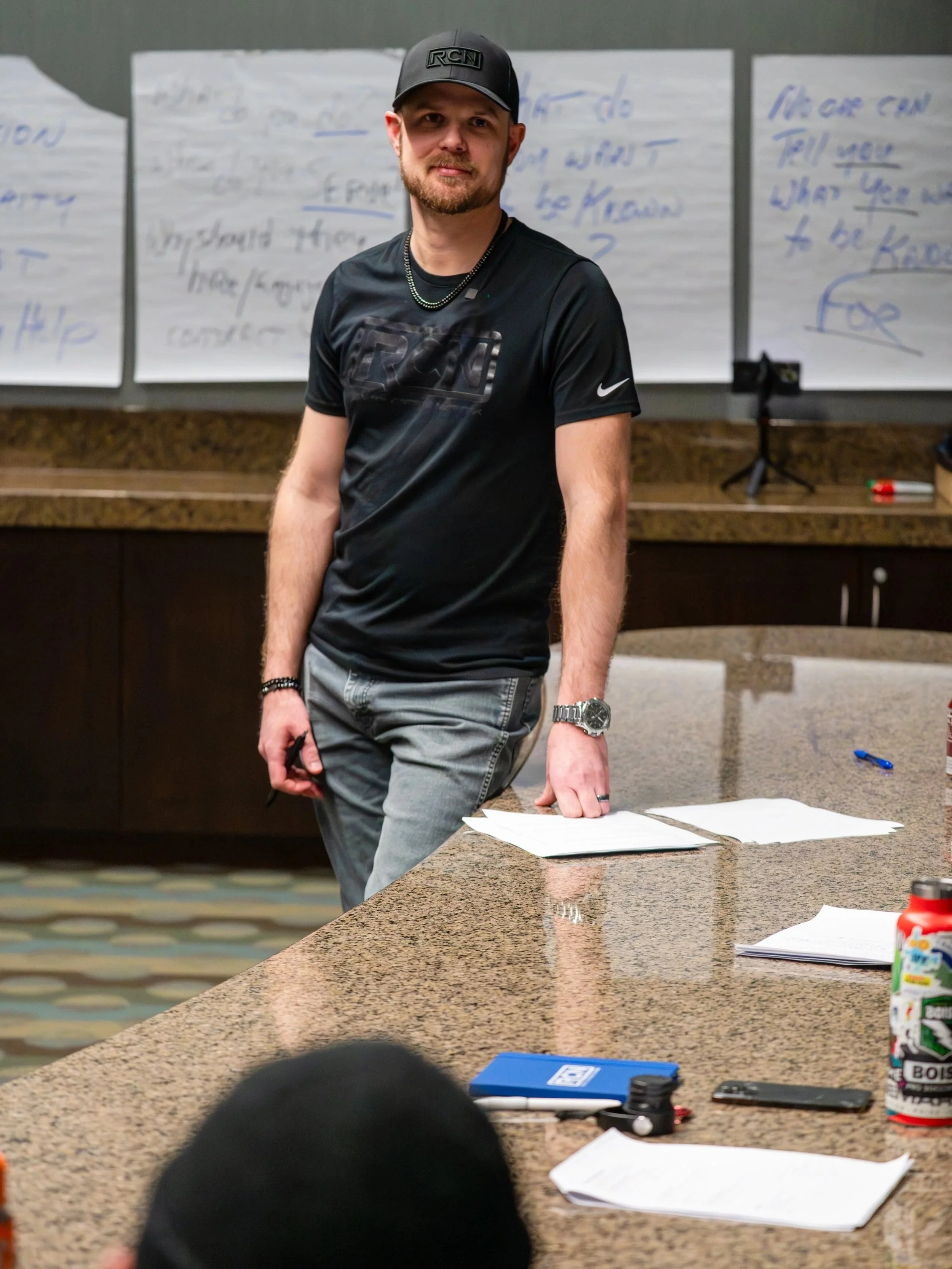 A man with a beard, wearing a black Nike T-shirt, a black cap, and a watch, standing in a conference room next to a granite table with papers, a blue notebook, and a water bottle. There are whiteboards with handwritten notes behind him.