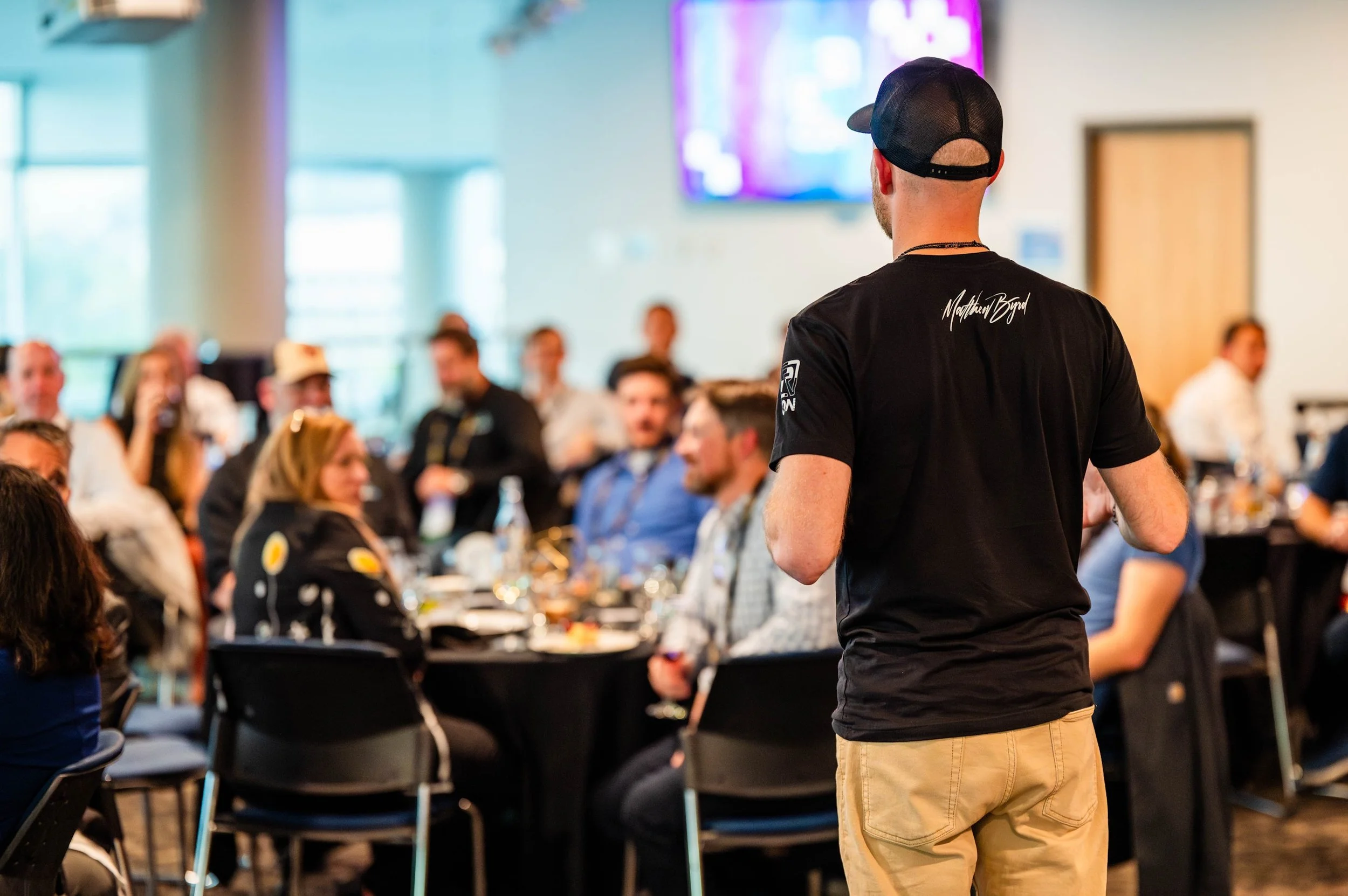 A man wearing a black t-shirt and beige pants facing an audience during a presentation or event at a large indoor space with seated attendees, tables, and a large screen in the background.