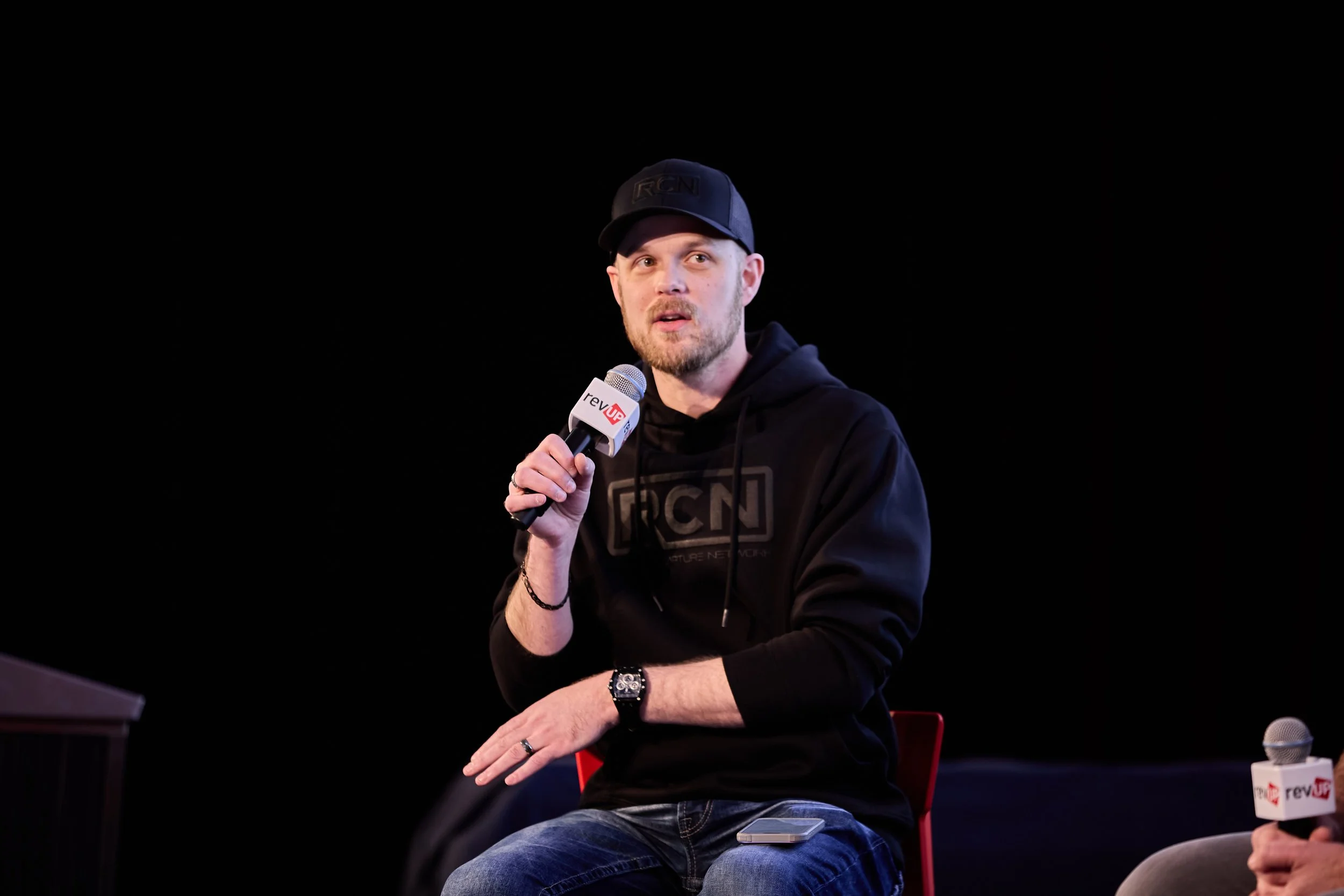 A man wearing a black hoodie with RCN logo, black cap, and jeans, sitting and holding a microphone with a revUP logo, speaking at an event with a dark background.