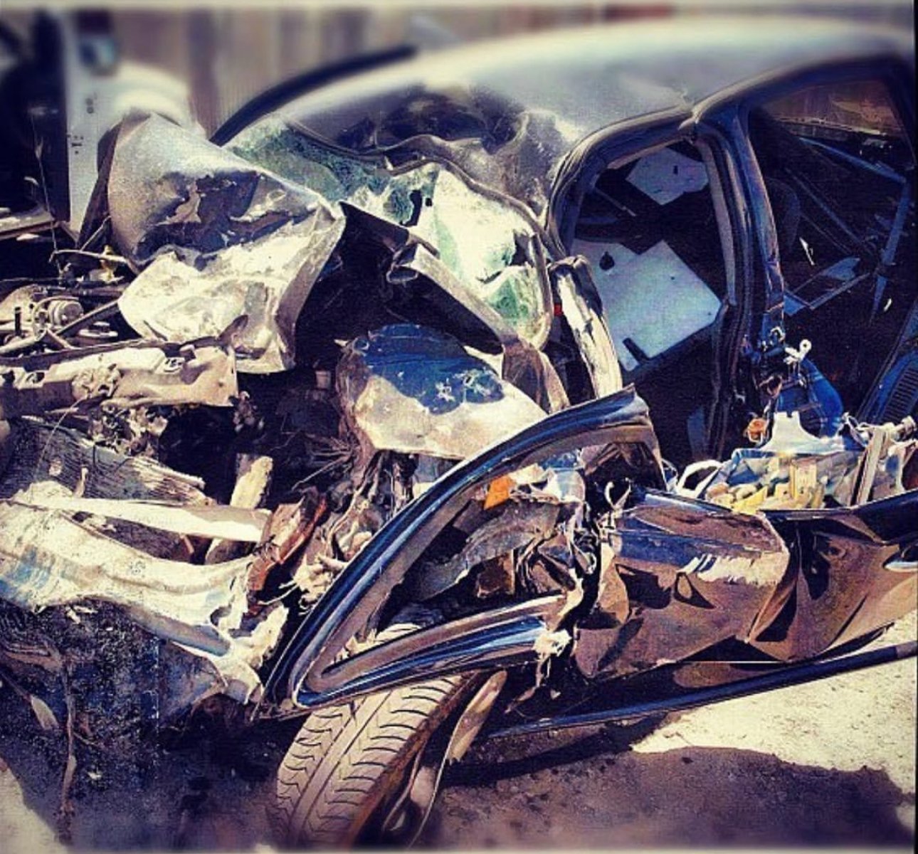 Damaged black car with extensive front-end crash, crumpled metal, shattered windshield, and deployed airbags.