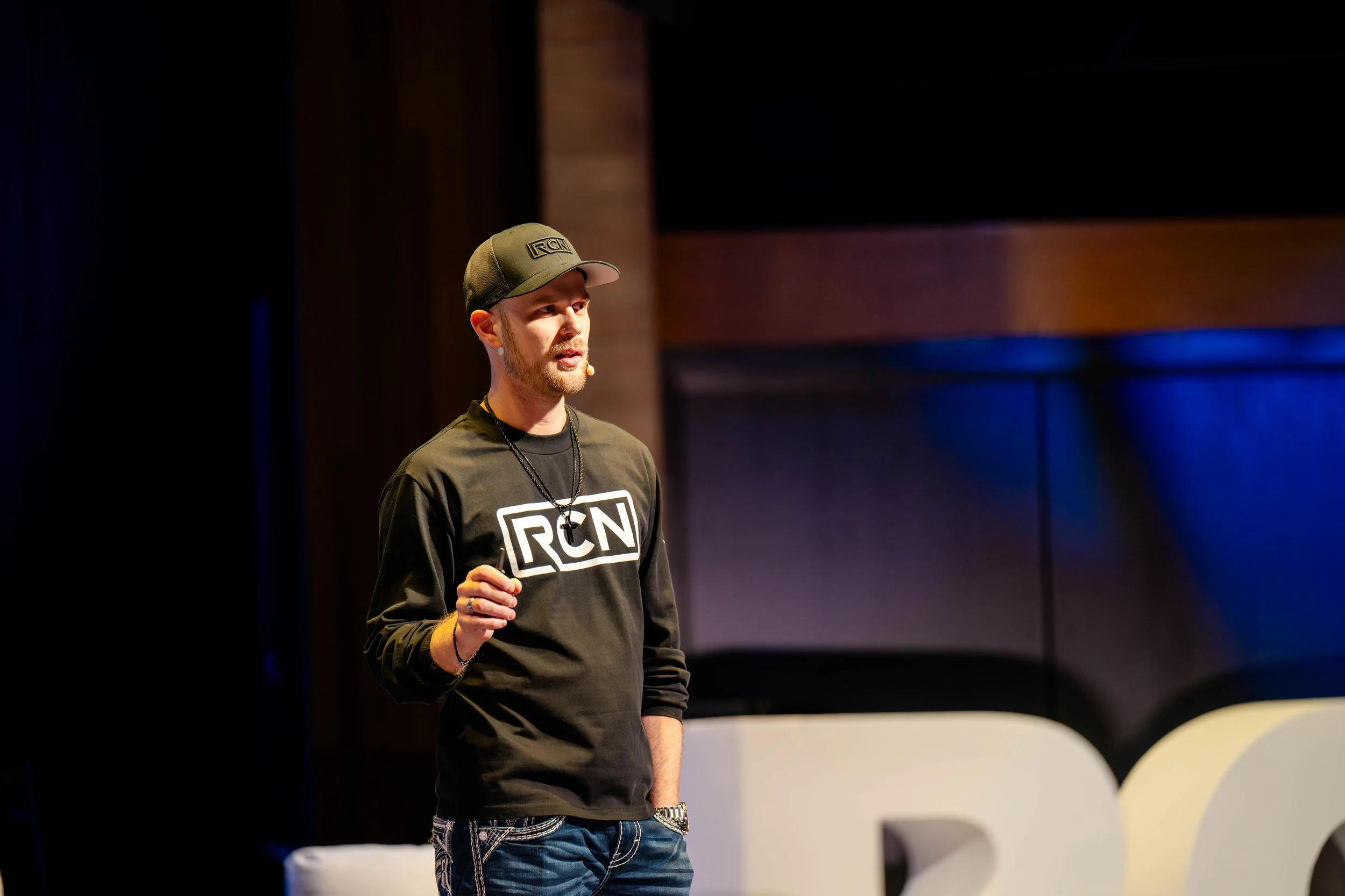 Young man speaking on stage, wearing a black RCNI sweatshirt and a baseball cap, with a wooden background and blue lighting.