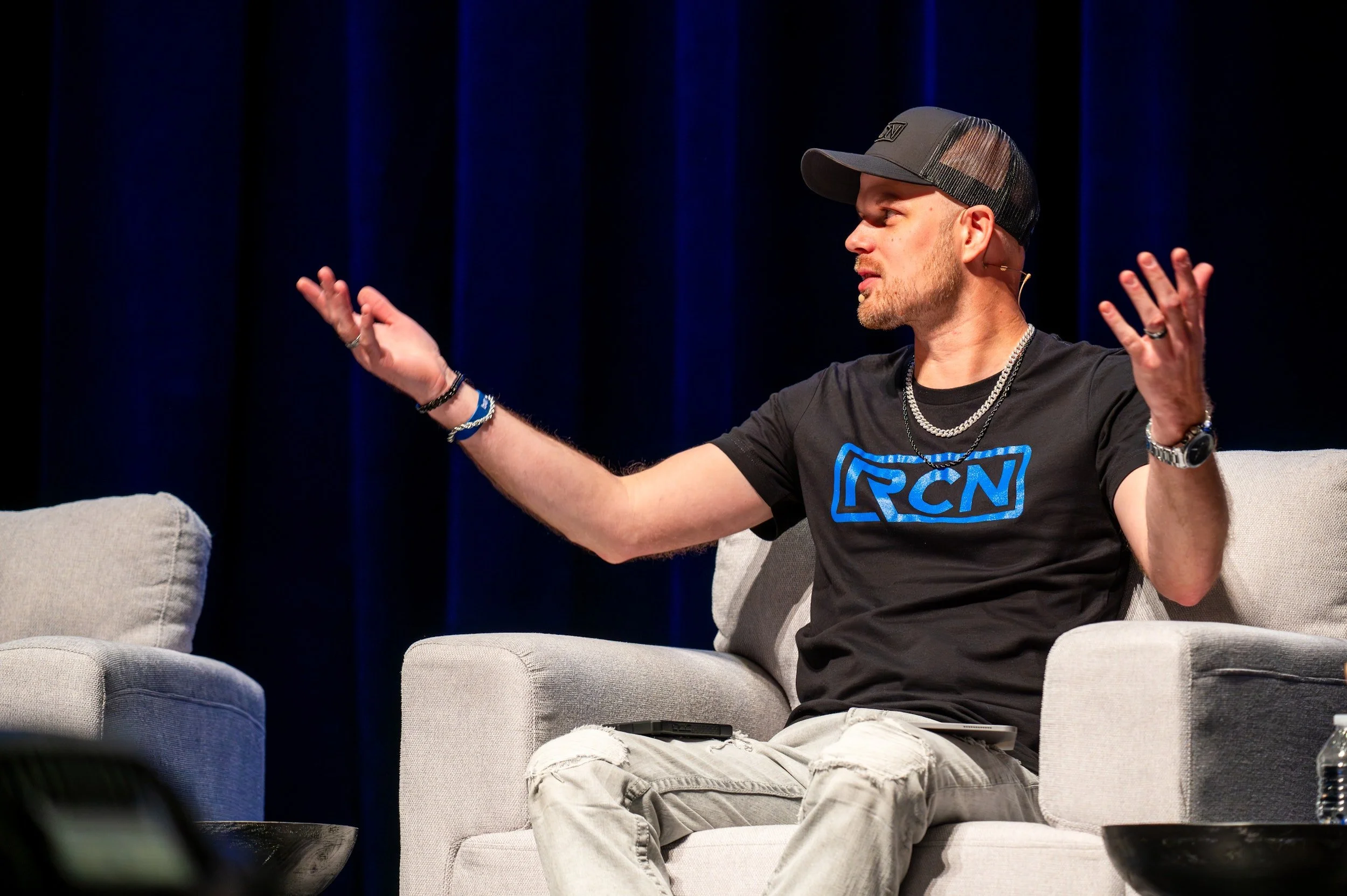 A man sitting on a gray sofa, gesturing with his hands, wearing a black RCW T-shirt, a dark baseball cap, and multiple necklaces, engaged in a conversation or presentation on stage with dark blue curtains in the background.