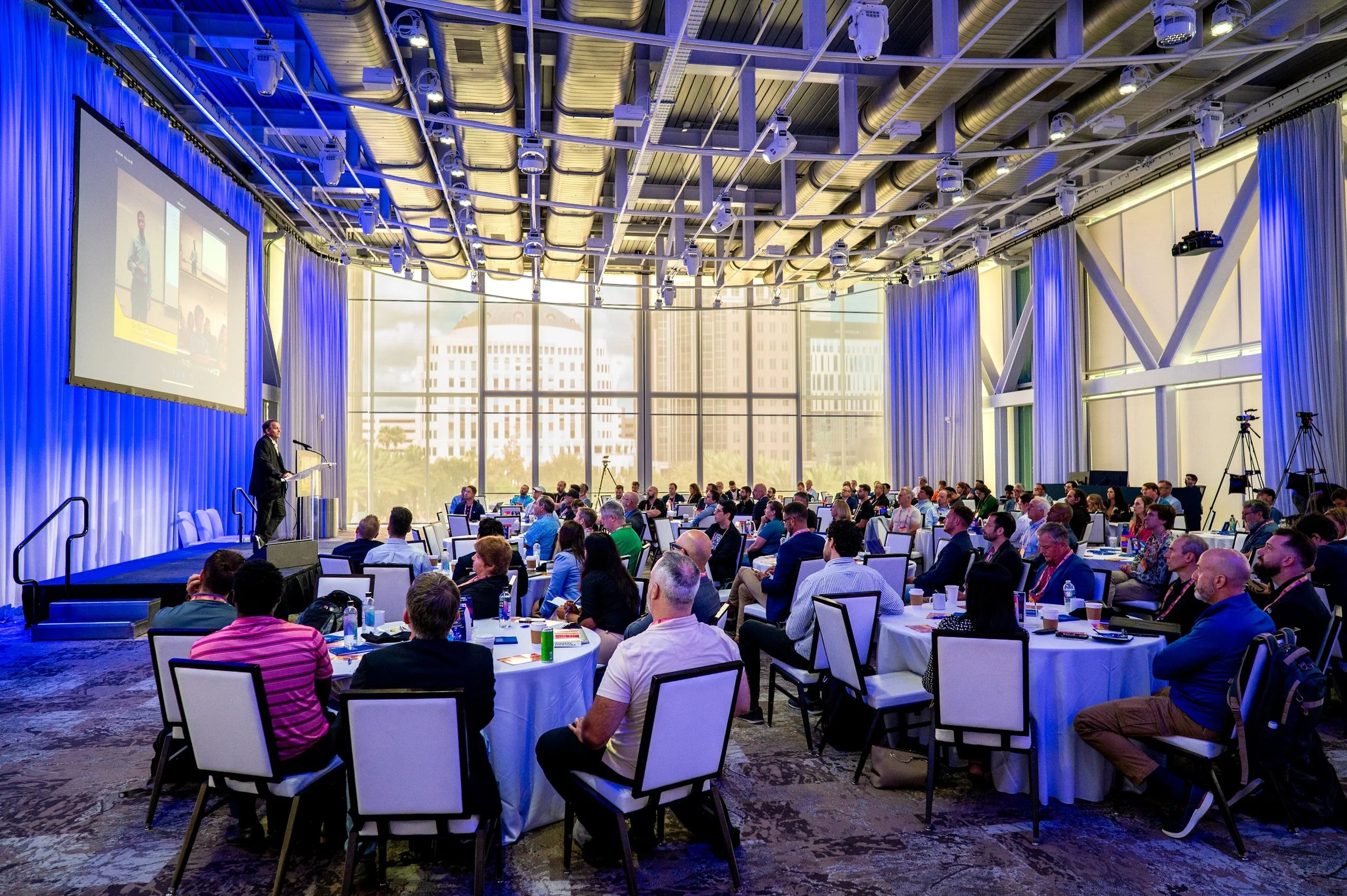 Conference room filled with seated attendees, a speaker on stage, large window with city view, multiple cameras, and a large screen projecting a presentation.
