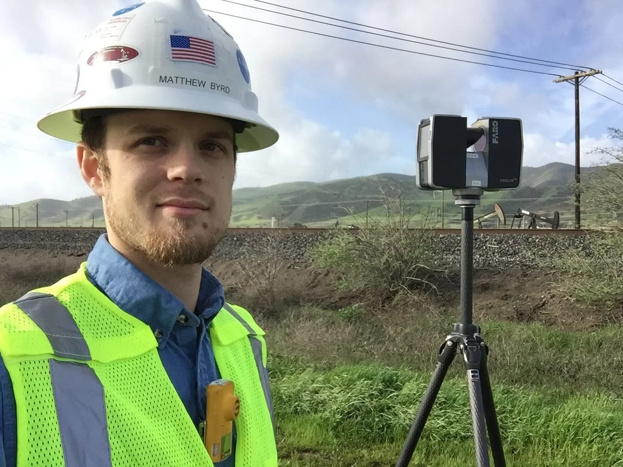 A man wearing a white construction helmet with an American flag sticker, a high-visibility vest, and a blue shirt stands outdoors near a tripod with a 3D laser scanner, with green hills and train tracks in the background.