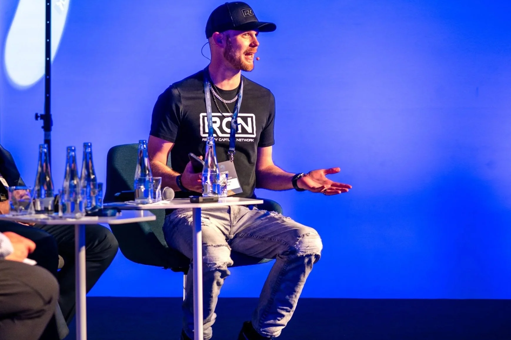 A man wearing a black RCNN t-shirt, a black cap, and ripped jeans is sitting on a chair, speaking at an event. There are several glass bottles of water on a small table in front of him, and he is gesturing with his right hand while holding a phone in his left hand. The background is blue.
