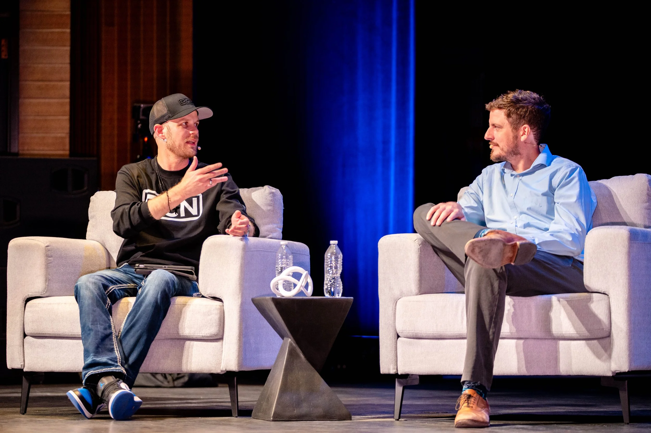 Two men sitting on white chairs having a conversation on stage. One man is wearing a black hoodie, jeans, and a baseball cap, gesturing with his hands. The other man is wearing a light blue shirt and dark pants, listening attentively. There are water bottles and decorative objects on a small table between them, with a dark background and stage lighting.