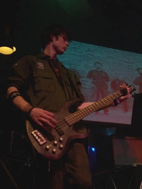 A young man playing a bass guitar on stage with a video screen behind him showing a group of people.