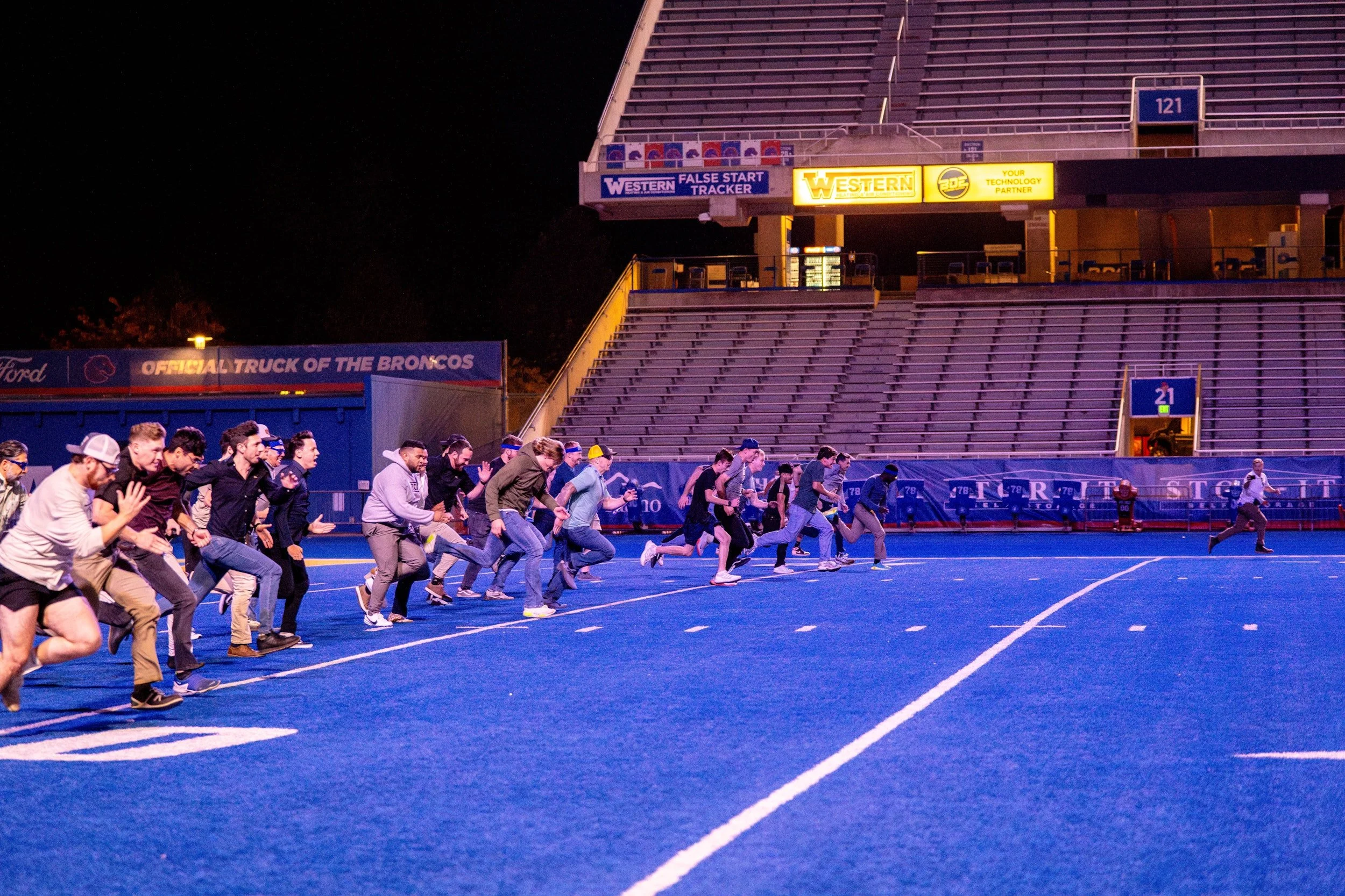 A group of people in casual attire running on a football field at night, with stadium seats and signs in the background.