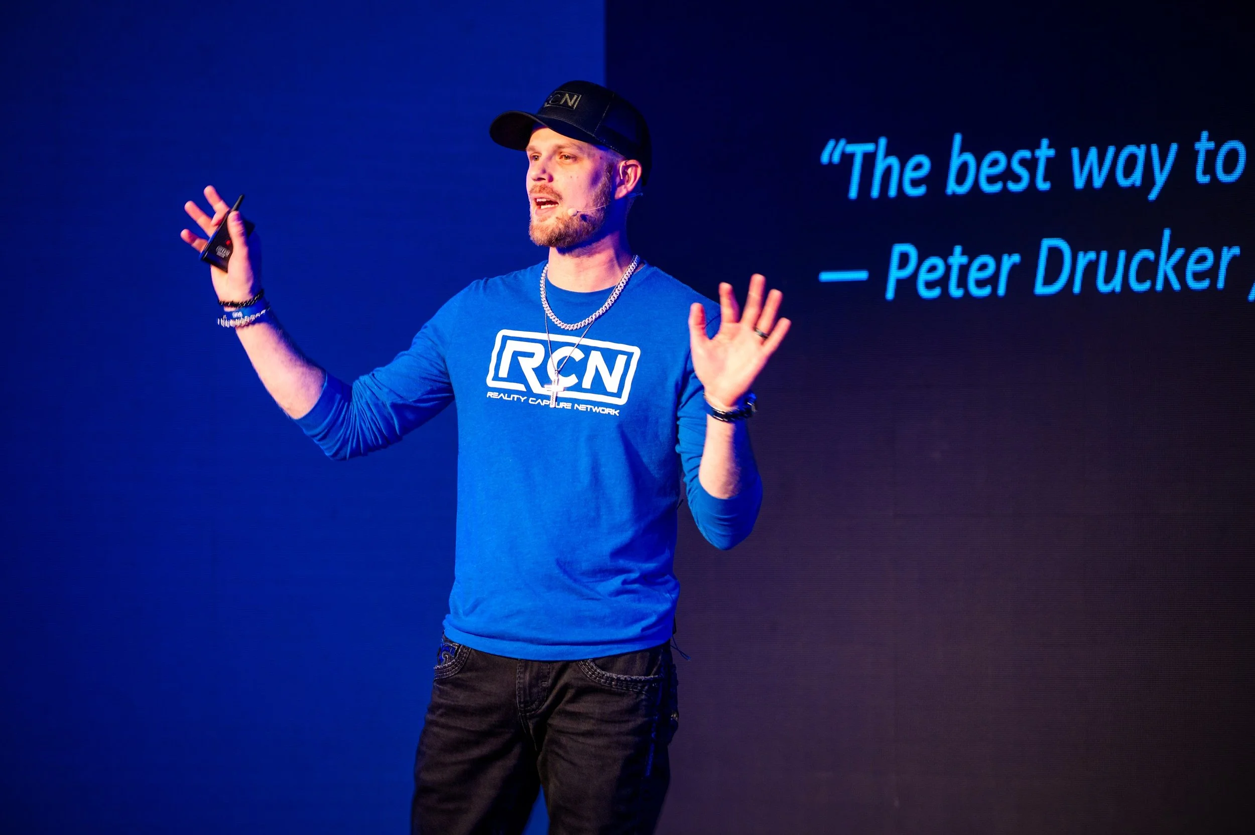 Man giving a presentation in front of a screen displaying a quote by Peter Drucker, wearing a blue RCN t-shirt, black cap, and accessories.