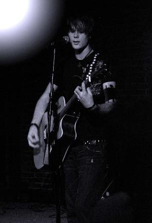 A young man playing an acoustic guitar and singing into a microphone on stage, with a spotlight overhead and a dark background.