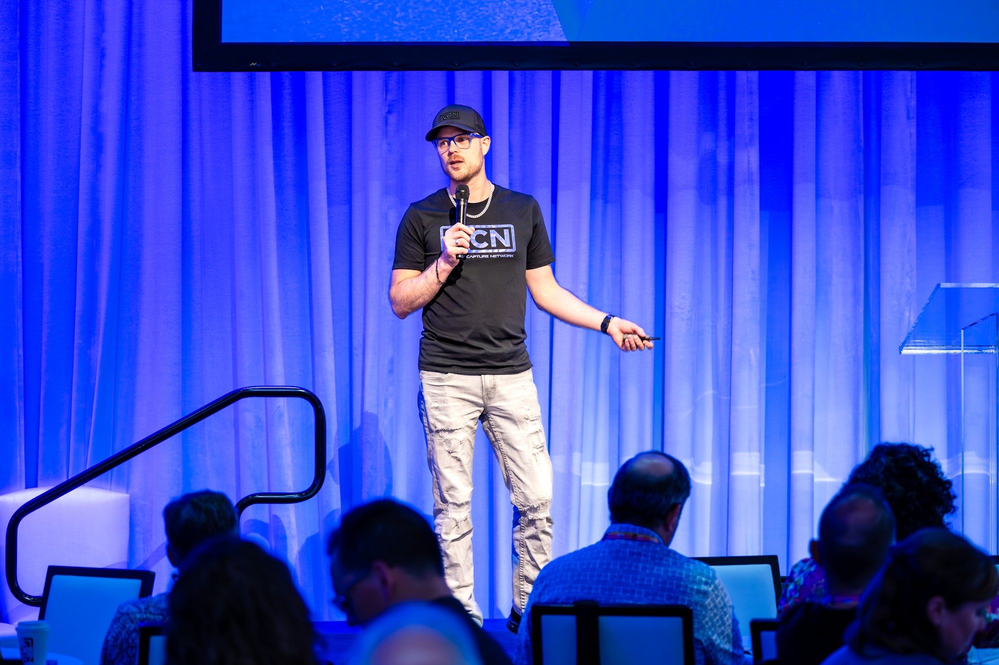 A man wearing a black T-shirt, beige ripped pants, glasses, and a black cap is speaking into a microphone on stage during a conference or presentation. He is holding a small remote in his other hand, with an audience seated in front of him.