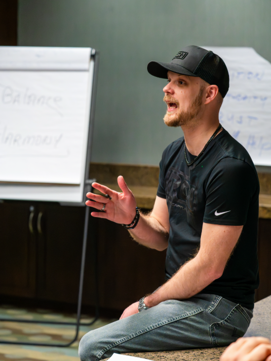A man wearing a black cap and black Nike t-shirt, sitting and speaking in a room with whiteboard and flip chart in the background.