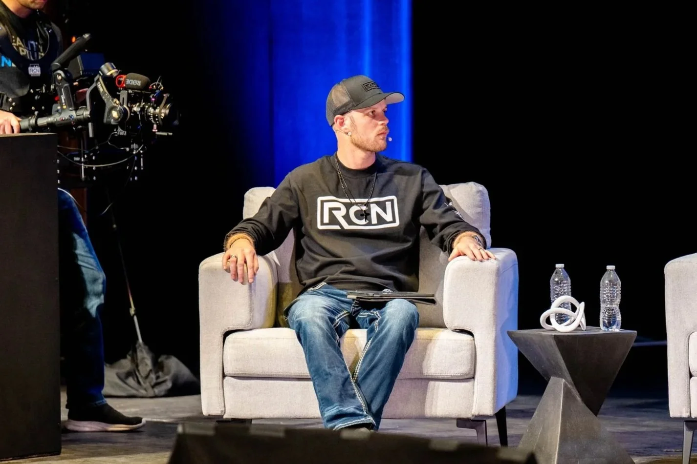 A man sitting on a beige armchair on a stage, wearing a black RCN sweatshirt and baseball cap, with a clip microphone, beside a small black table with two water bottles and headphones, with blue lighting in the background.