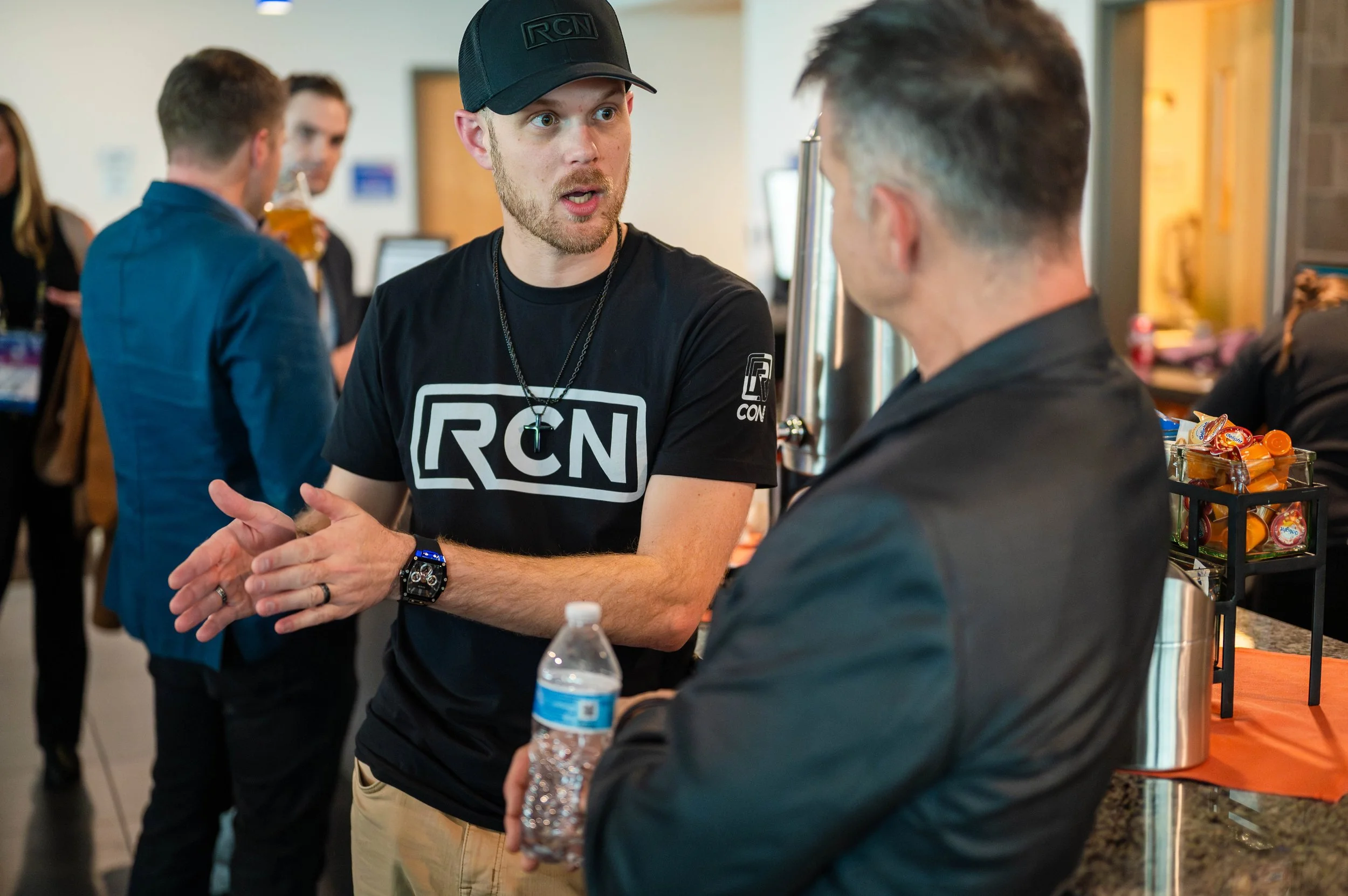 Two men are engaged in a conversation at an event, with one man wearing a RCN t-shirt and a black cap, and the other man holding a water bottle. In the background, other people are socializing, with one person drinking a beverage and others near a table with snacks.
