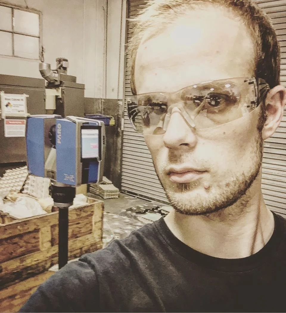 A man wearing safety goggles takes a selfie in a workshop with machinery and wooden crates in the background.
