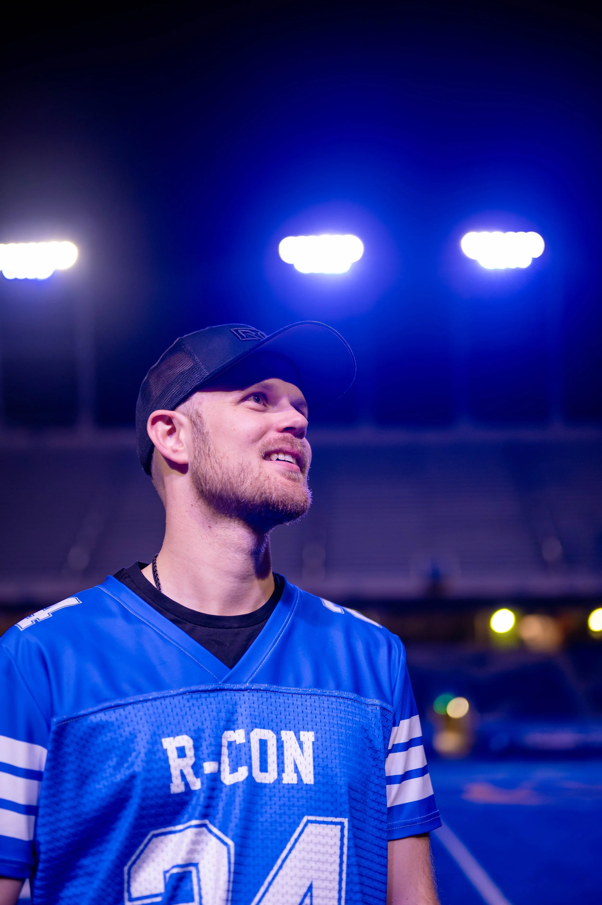 A young man wearing a blue football jersey with 'R-CON' and the number 24, a black cap, and a black shirt underneath, stands on a football field at night under bright stadium lights, smiling and looking upward.