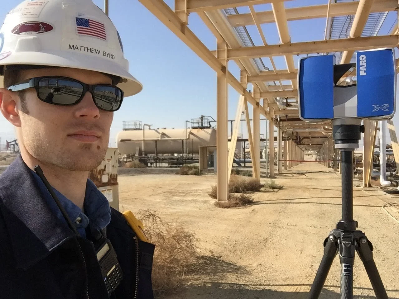 A man wearing a white hard hat with an American flag and name tag 'Matthew Byrd', sunglasses, and a blue shirt, standing next to a blue environmental monitoring instrument on a tripod in an industrial outdoor area with pipelines and structures.