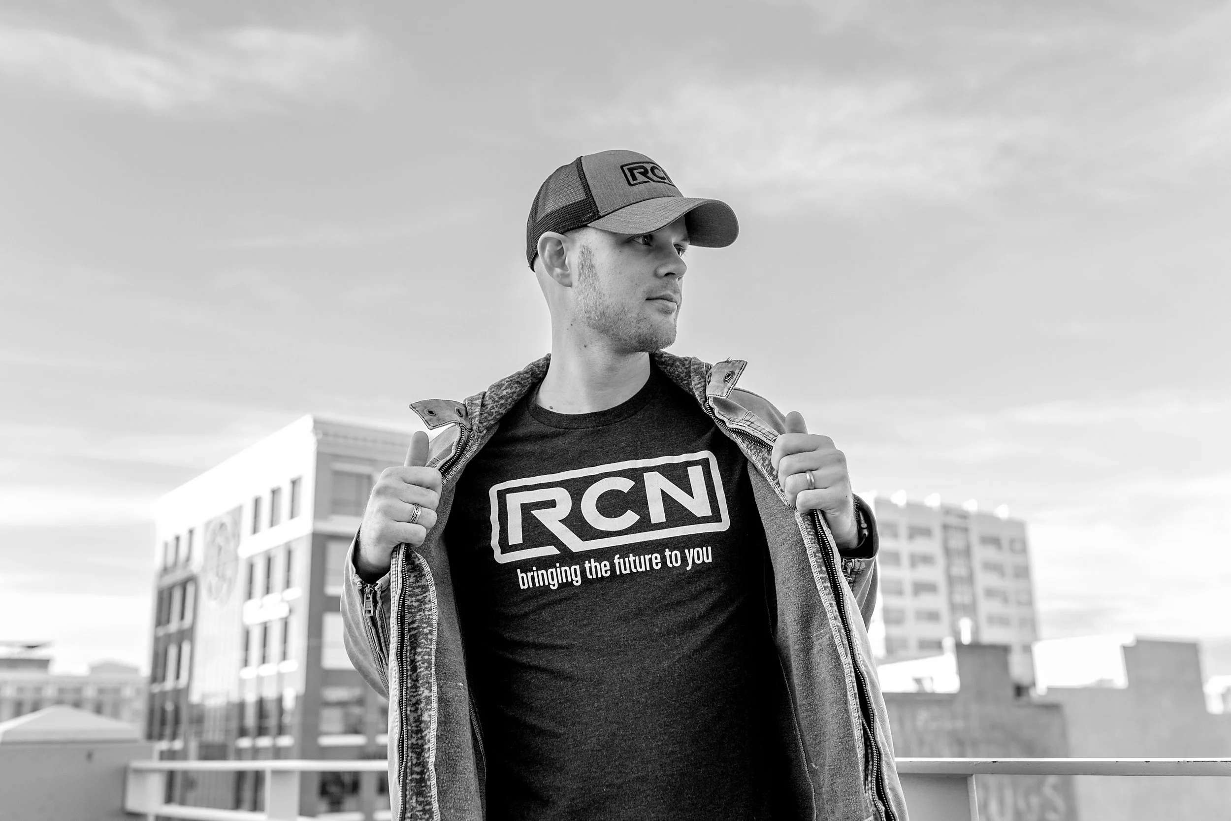 A man in a cap and jacket revealing a T-shirt with 'RCN' and 'bringing the future to you' on it, standing outdoors with buildings in the background.
