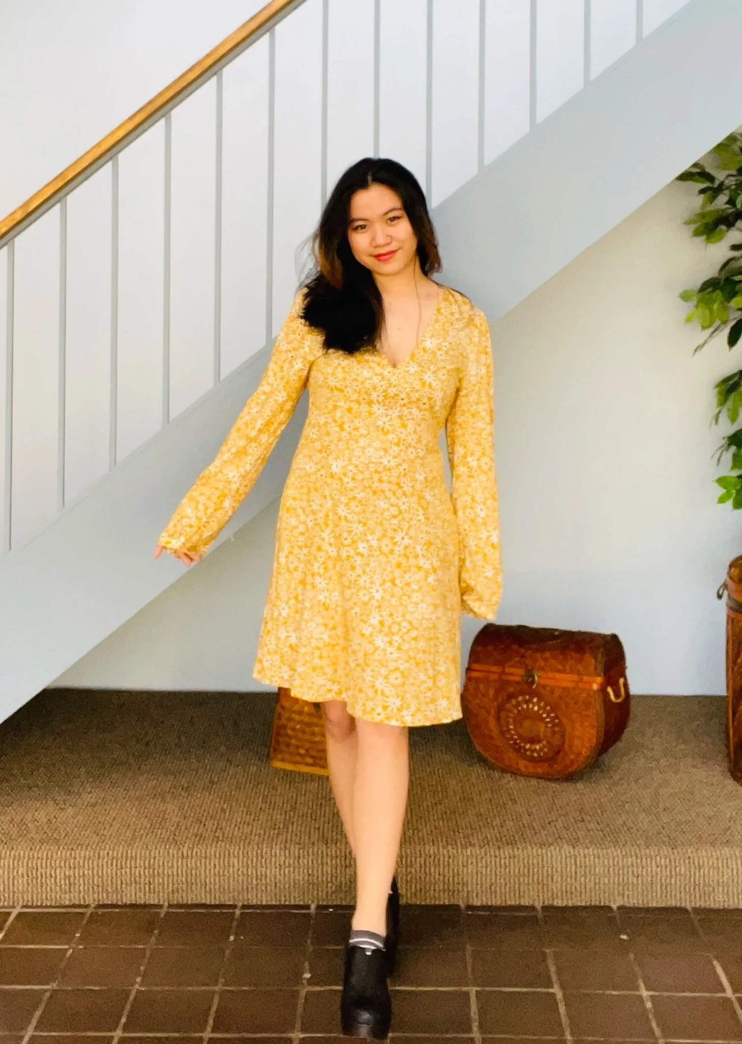 A woman in a yellow floral dress standing in front of a staircase and a white wall, with a green plant and a decorative brown chest nearby.