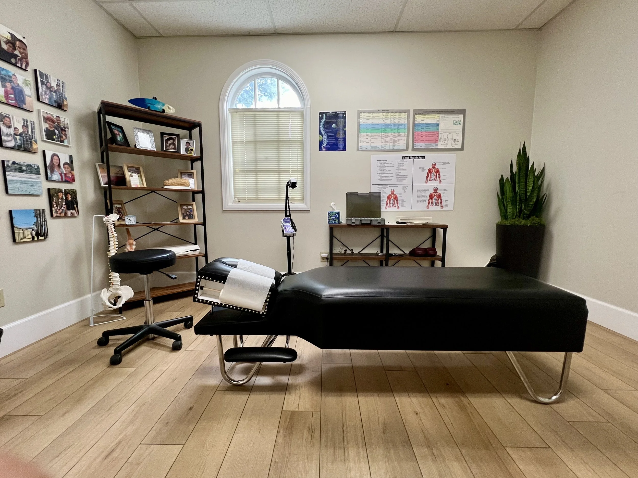 Medical examination room with a black examination table, a rolling stool, a computer desk, and medical posters on the wall. There is a window with blinds, a tall potted plant, and a shelf with photos and decor.