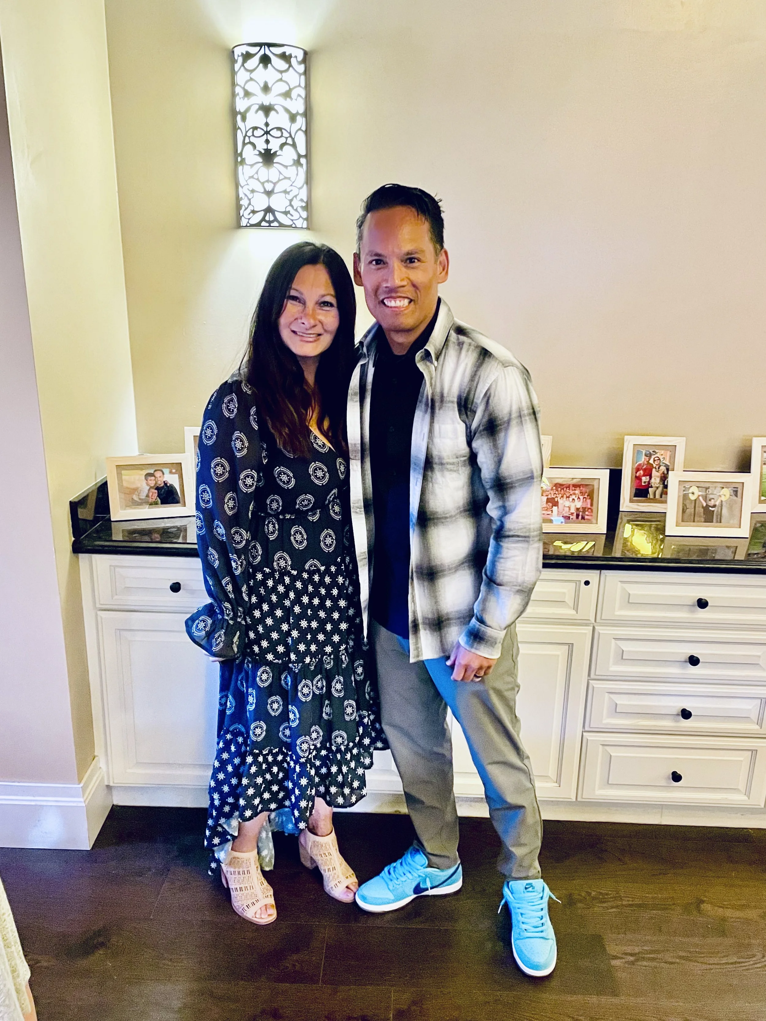 A man and woman standing together indoors, smiling at the camera. Behind them is a white cabinet with framed photos and a decorative wall sconce.