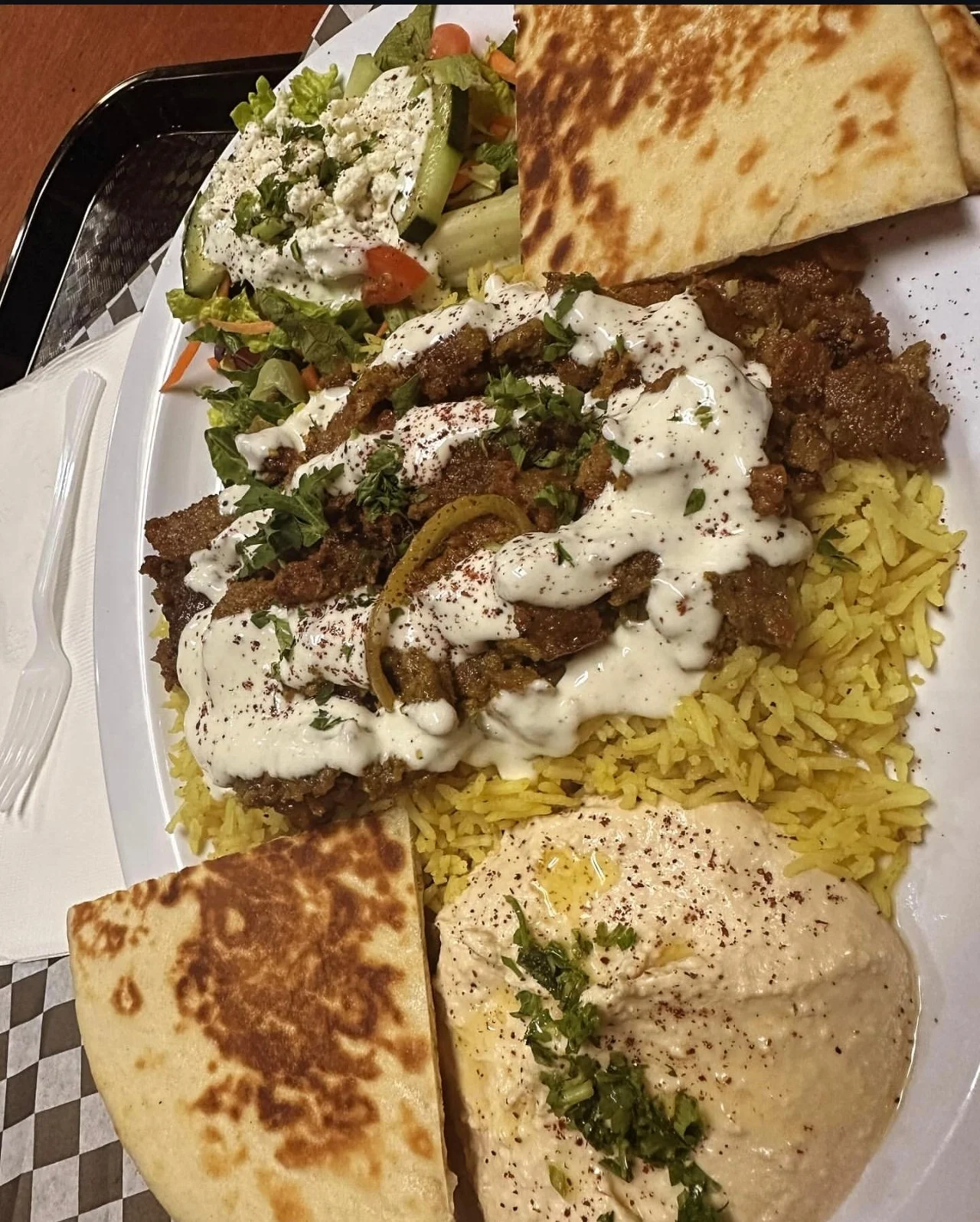 A plate of Middle Eastern and Mediterranean dishes including yellow rice, lamb with white sauce, hummus, fresh salad, and flatbread.