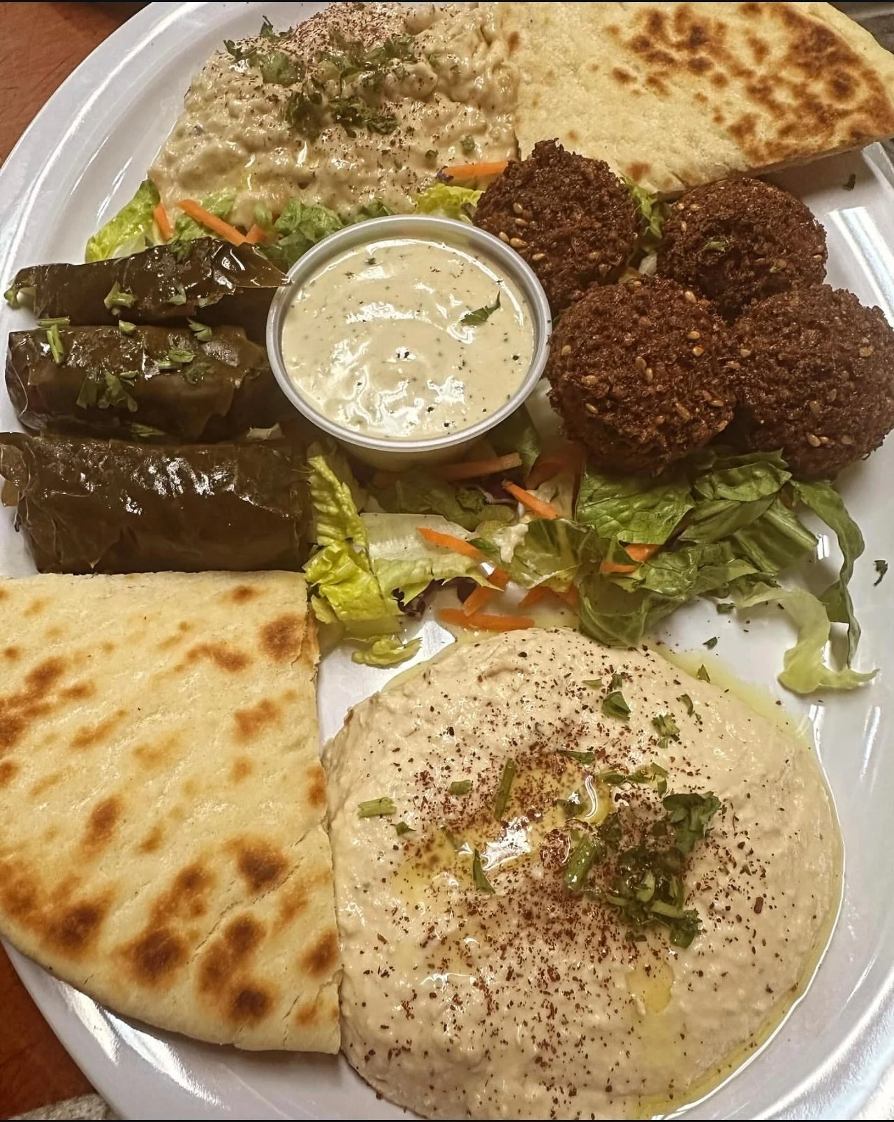 A plate of Middle Eastern food featuring hummus, falafel, stuffed grape leaves, flatbread, and a side of salad with cucumber, tomato, and lettuce, served with a dipping sauce.