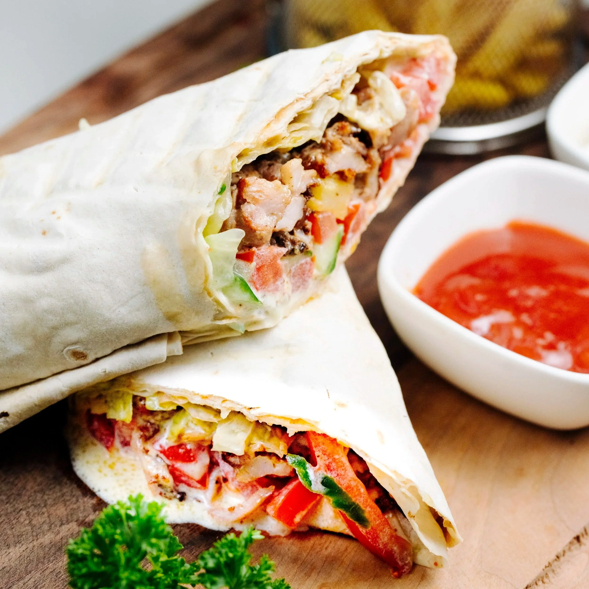Close-up of a wrap sandwich filled with meat, vegetables, and cheese, served with a side of parsley and bowl of red dipping sauce.