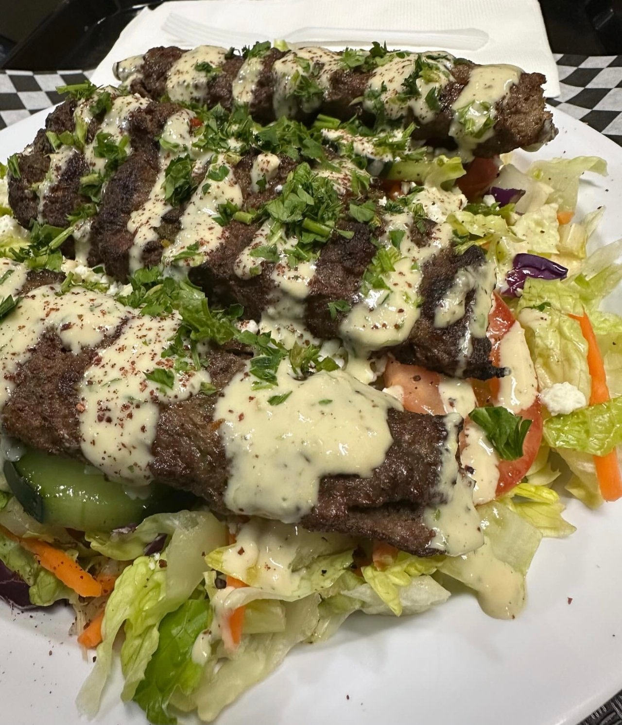 A plate of grilled beef ribs topped with creamy sauce and chopped cilantro, served over a bed of fresh salad with lettuce, tomatoes, cucumbers, and shredded carrots.