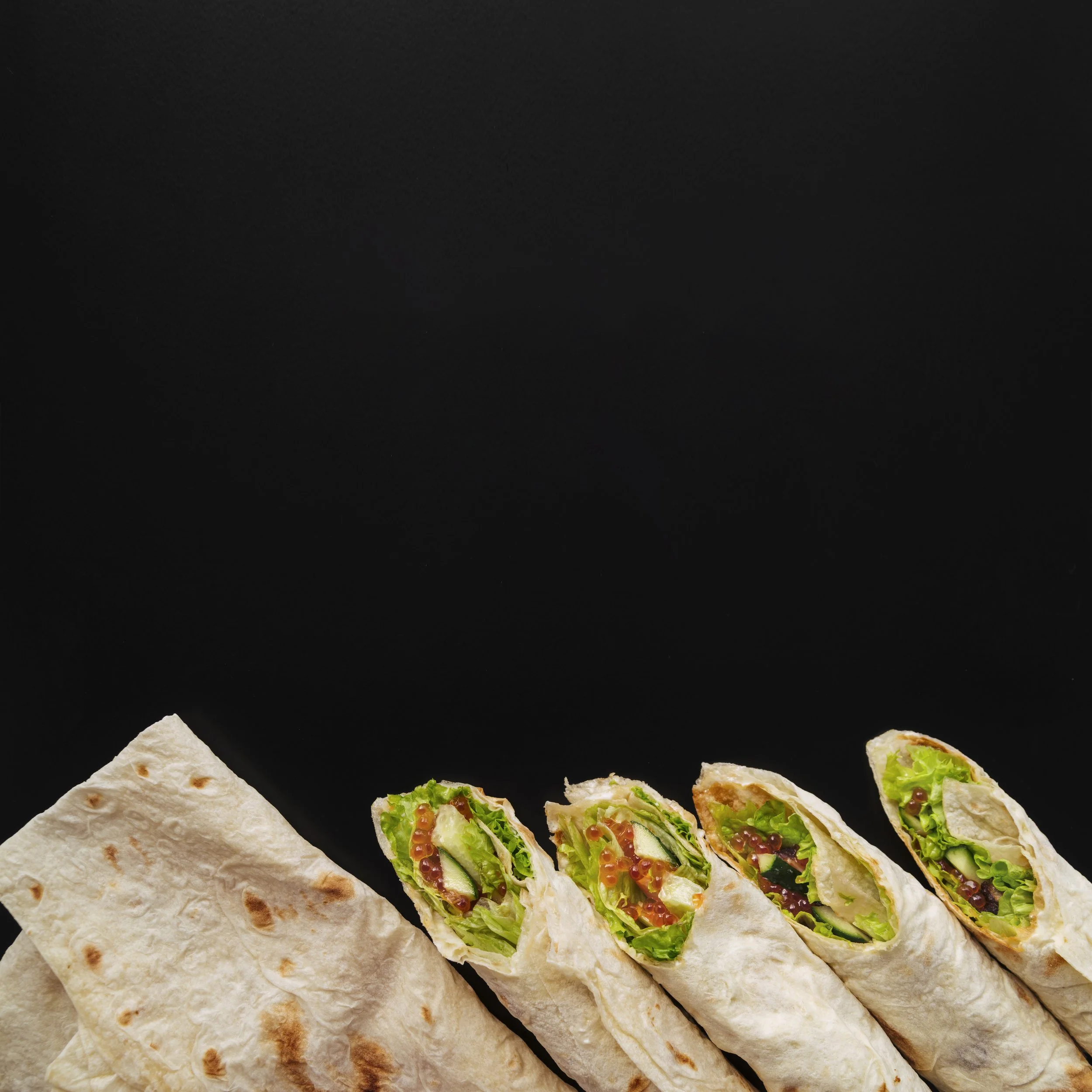 Close-up of three sandwich wraps filled with lettuce, cucumber, and smoked salmon or fish roe, with two triangular pieces of flatbread on a black background.