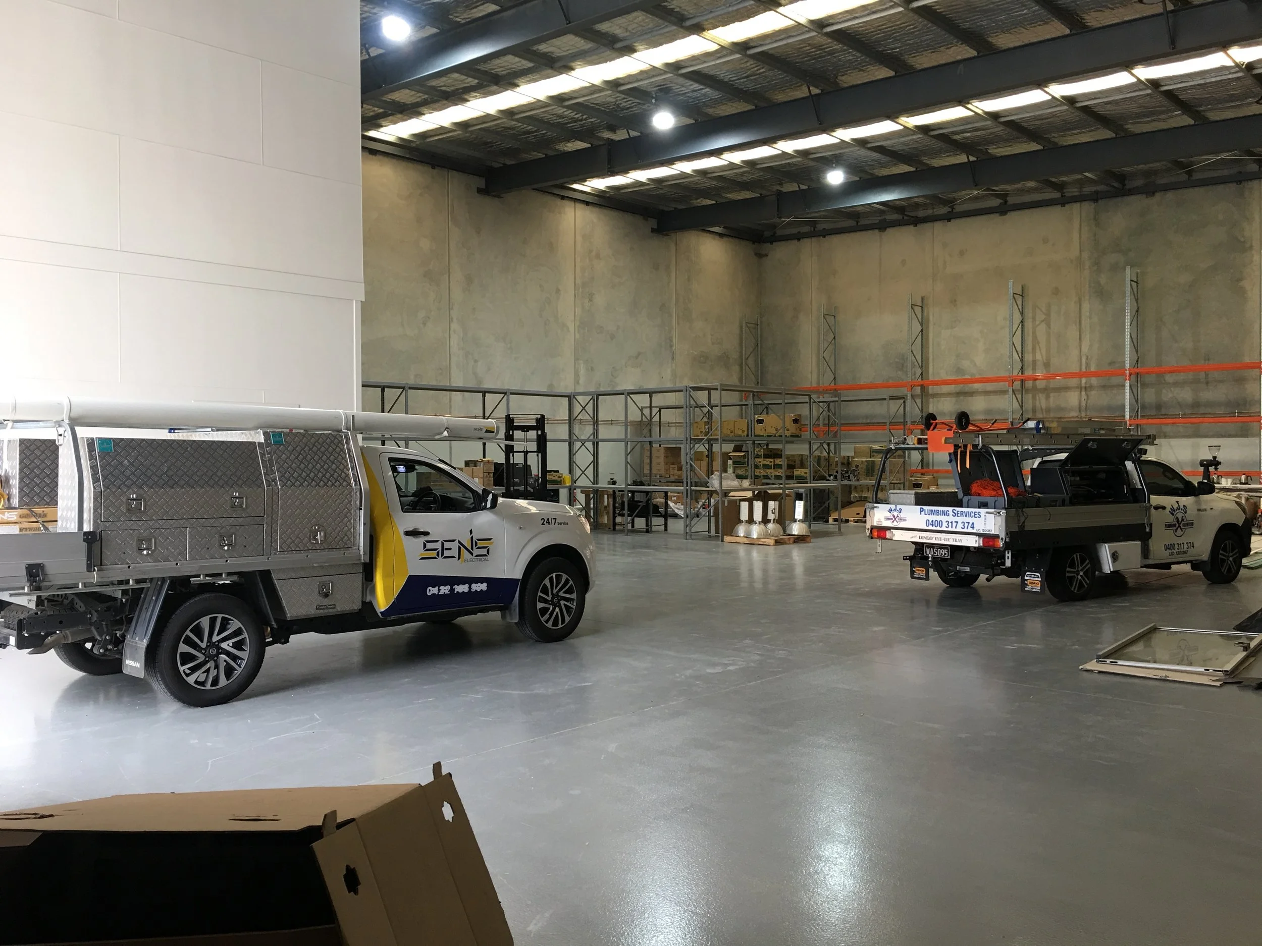 Two utility trucks parked inside a warehouse; the trucks have company logos and contact information, and the warehouse has metal shelving units and construction materials.