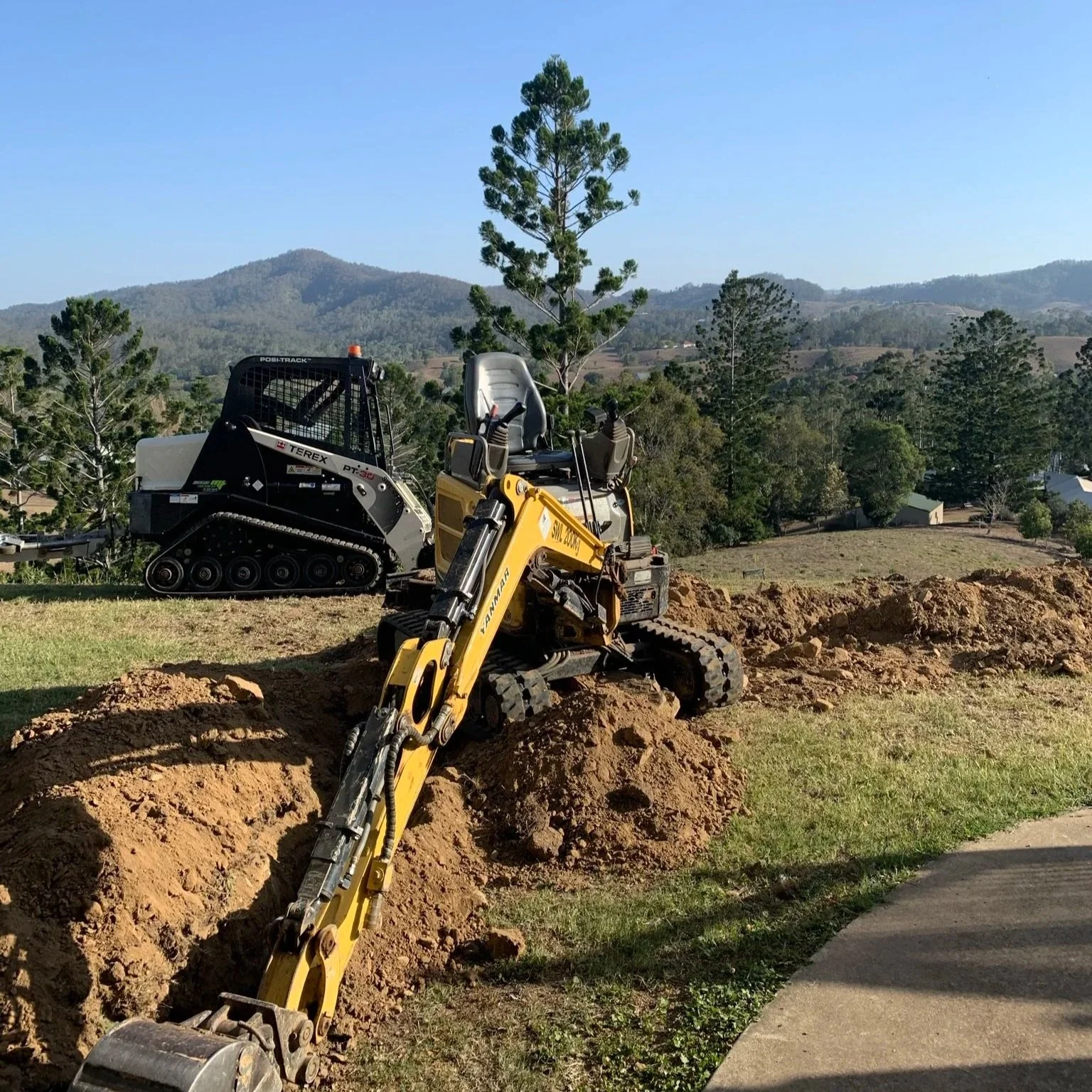 A yellow compact excavator digging a trench in a grassy area with trees and hills in the background.