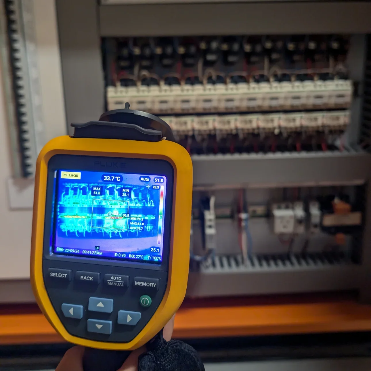 A thermal imaging camera held in front of an electrical panel with open circuit breakers and wiring inside.