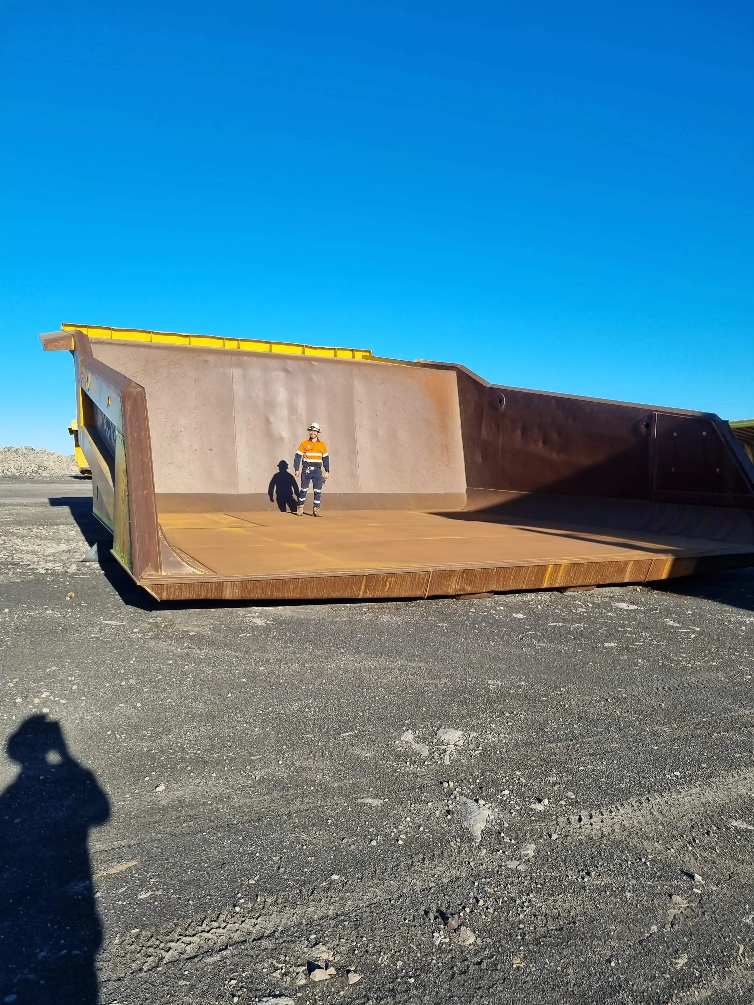 A construction worker in safety gear standing inside a large, empty haul truck tray on a construction site with a clear blue sky.
