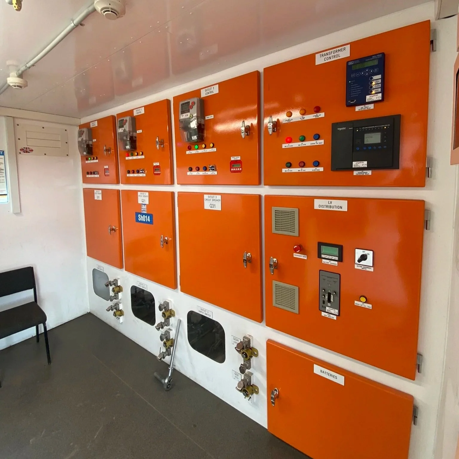 Electrical control panels on an orange wall with switches, meters, and labels, and a black chair nearby.