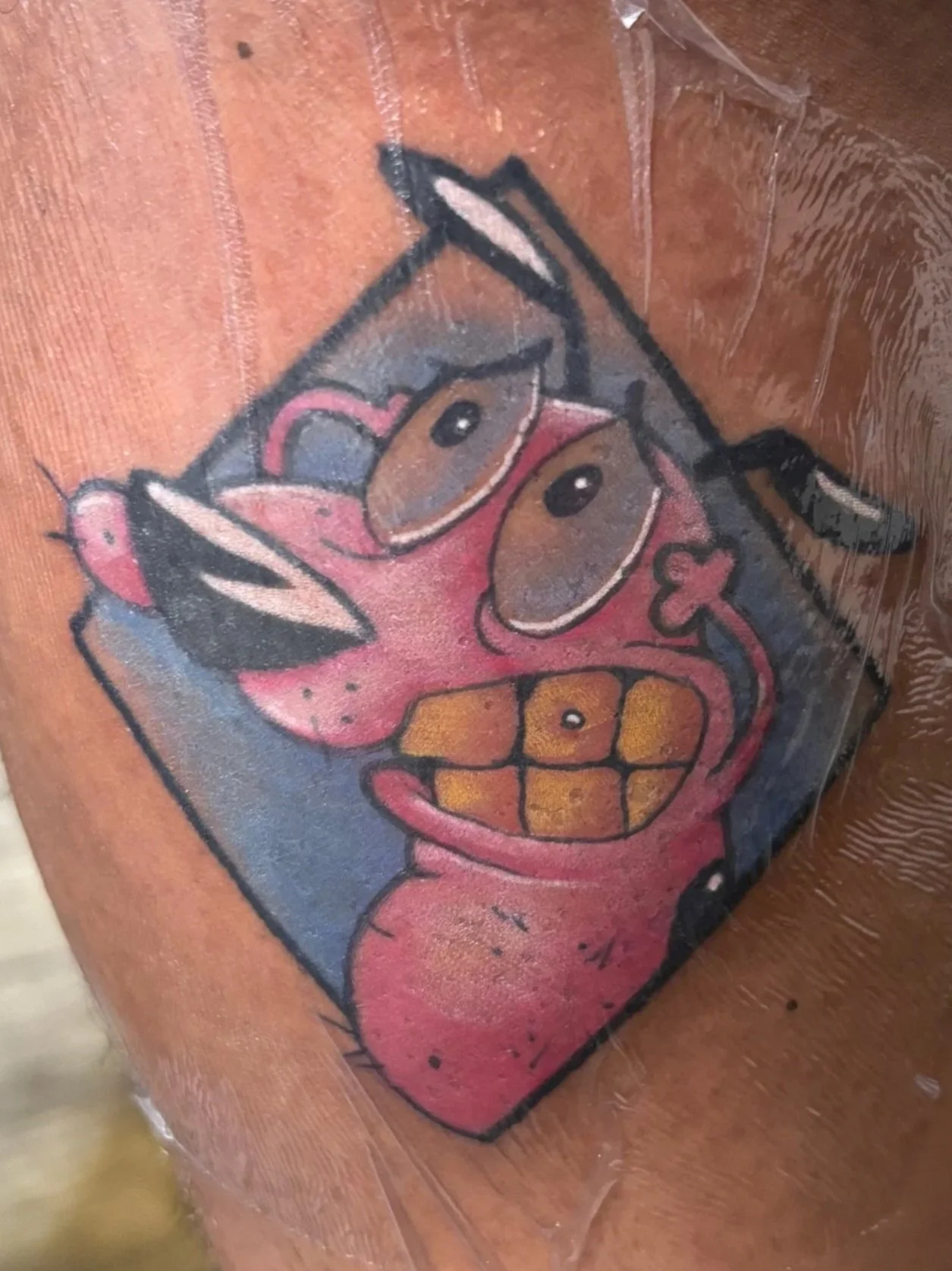 Tattoo of courage the cowardly dog with exaggerated features and a gold tooth, covered with clear plastic wrap on a person's leg.