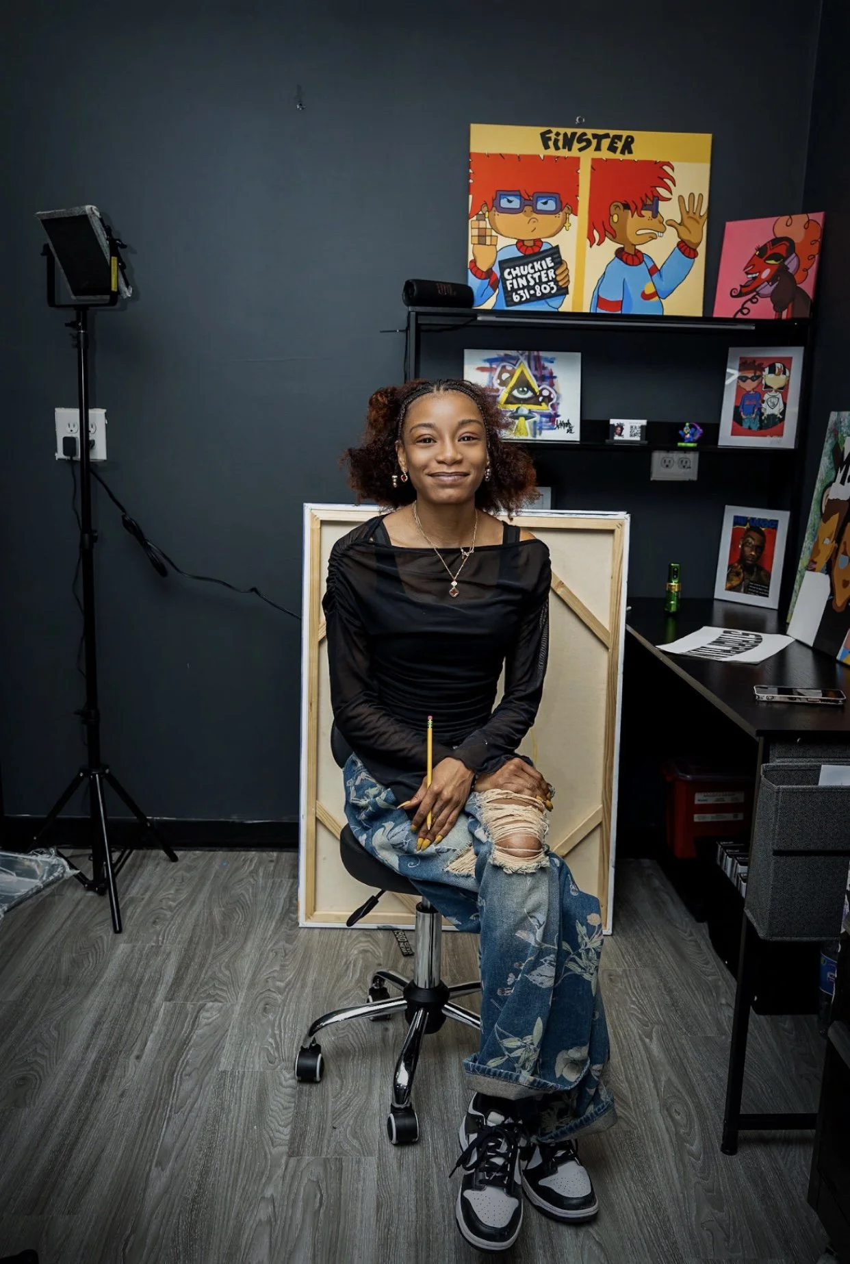Young woman sitting on a stool in a studio, holding a pencil, with colorful pop art posters behind her, wearing ripped jeans, a black sheer top, and black and white sneakers.