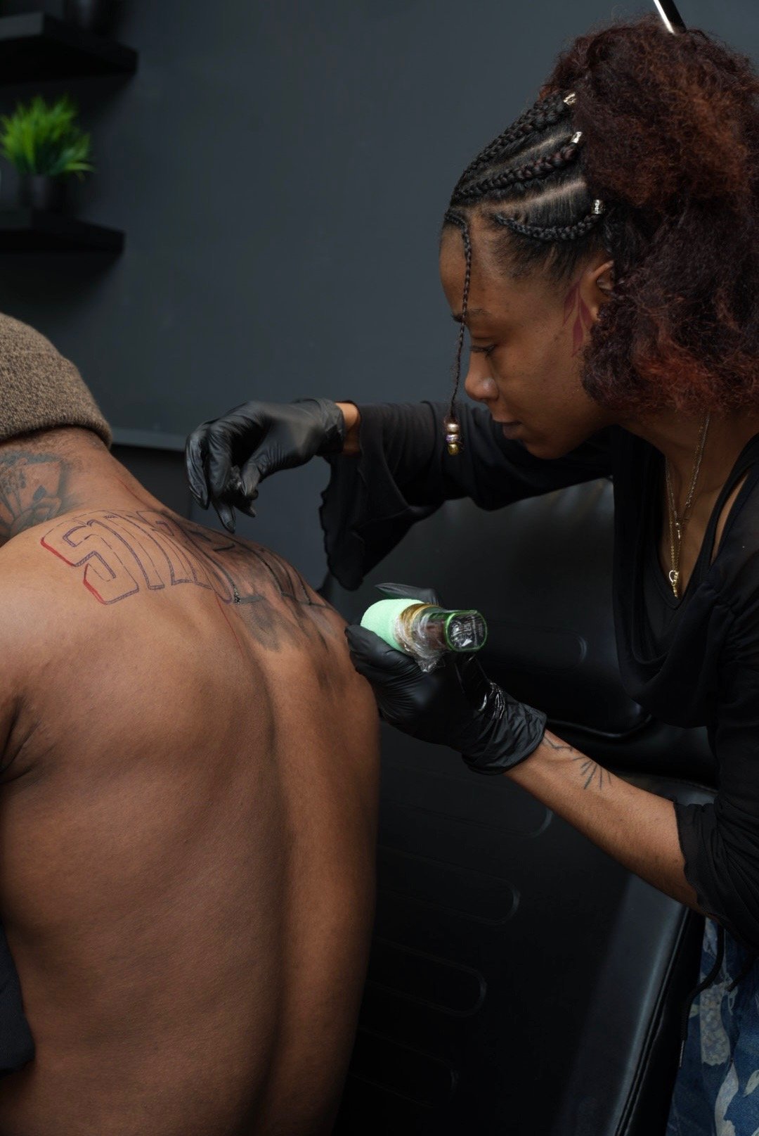 A tattoo artist is creating a design on a man's upper back with a tattoo machine, while the man is shirtless and the artist is wearing black gloves and a black shirt.