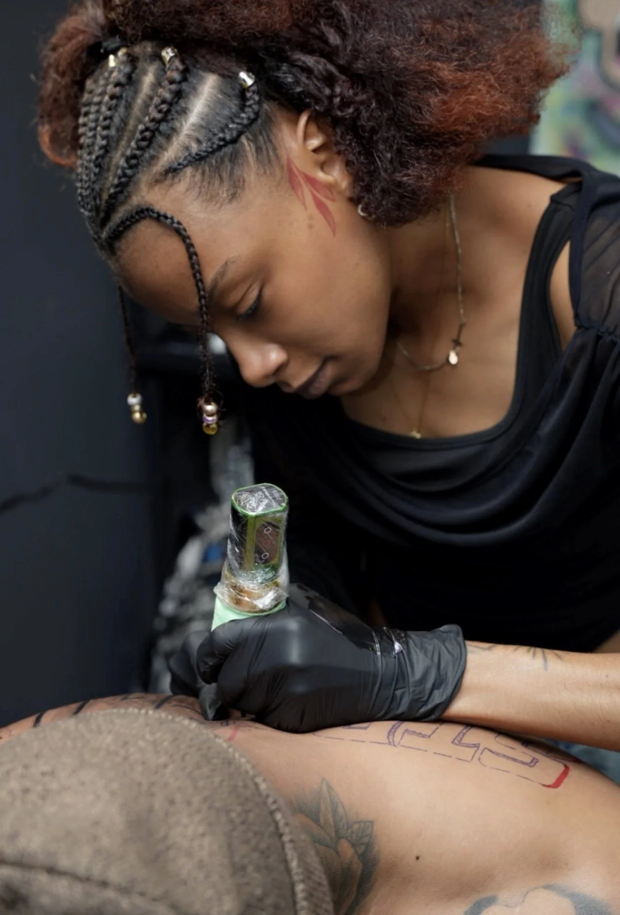A woman with braided hair and tattoos is tattooing a person's arm, who is lying down. The woman is holding a tattoo machine wrapped in plastic, and she is wearing gloves.