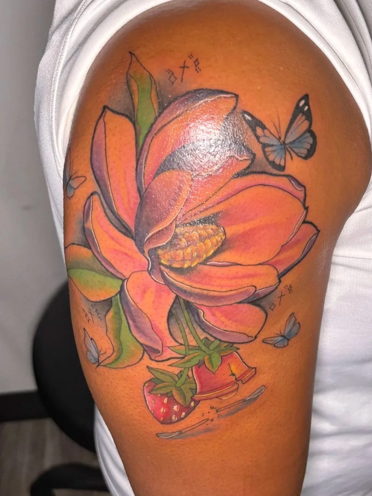Colorful tattoo of a blooming pink flower with green leaves on the woman's upper arm, accompanied by butterflies and small decorative elements.