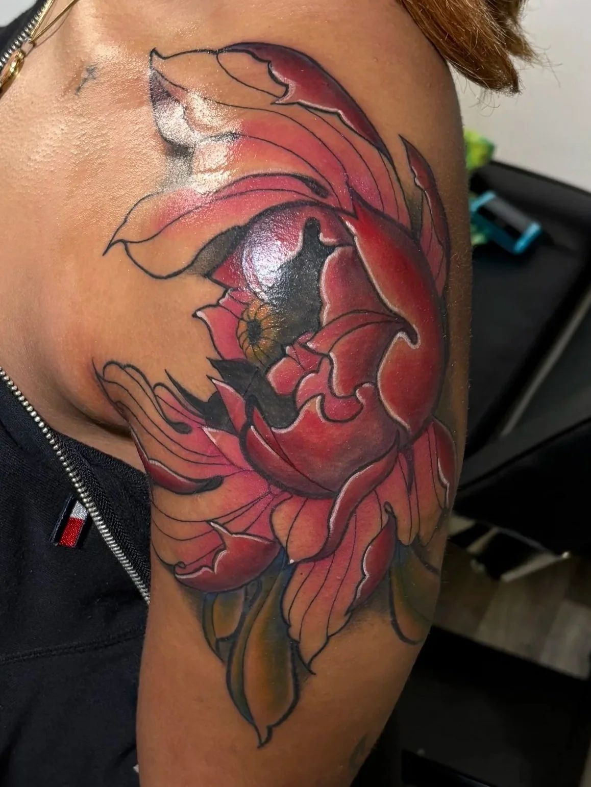 A woman with shoulder-length reddish hair showing a colorful floral tattoo of a large flower on her upper arm and shoulder. She is wearing a black sleeveless top, black pants, and turquoise shoes, standing in a room with wooden floors and a black massage or treatment table.